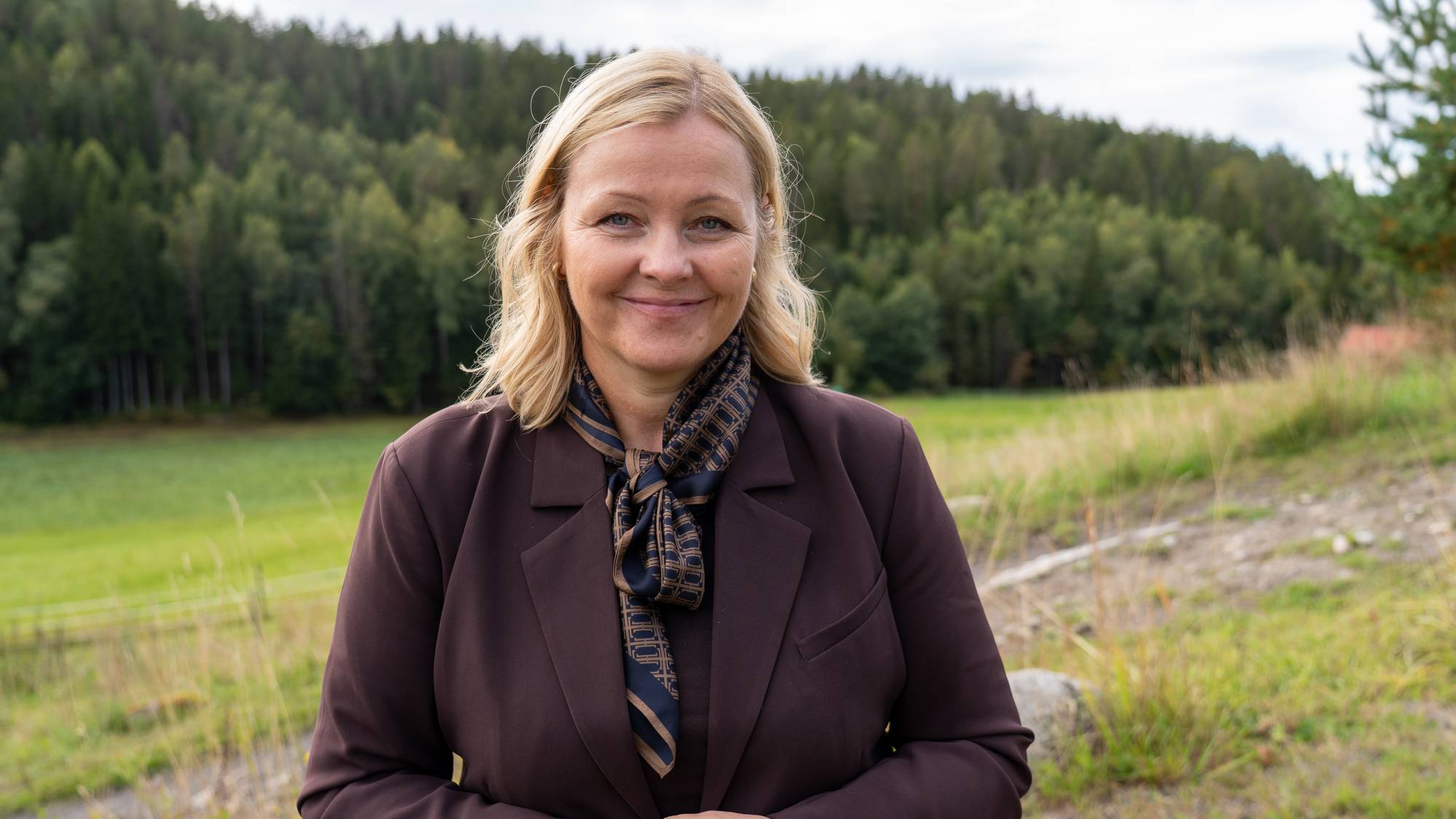 Kvinne med lyst hår, mørk dressjakke og skjerf, avbildet utendørs foran et skoglandskap. (Bildebeskrivelsen er laget av en KI-tjeneste)