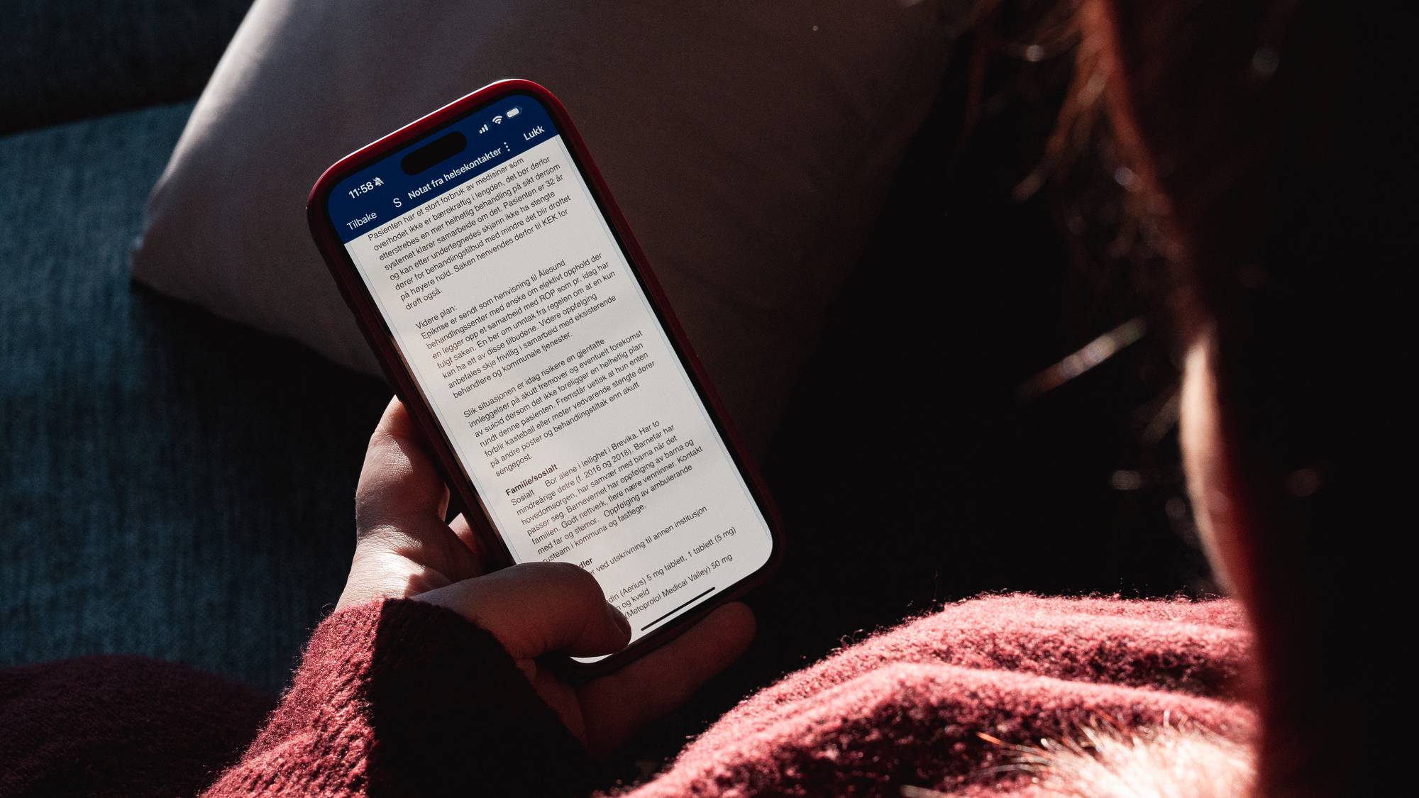 En person sitter i en sofa og holder en smarttelefon i hånden. Skjermen viser tekst, muligens fra en nettside eller et dokument. Omgivelsene er svakt opplyst, med en pute synlig i bakgrunnen. Personen har på seg en rød genser. (Bildebeskrivelsen er laget av en KI-tjeneste)