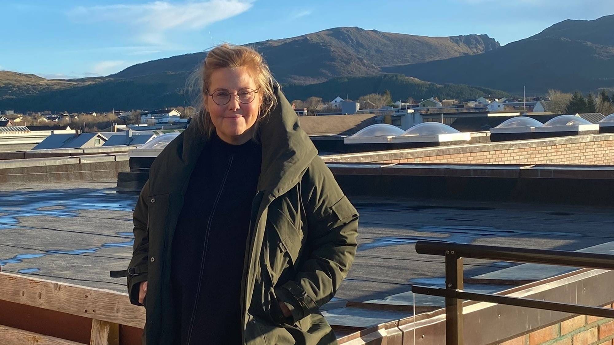 Kvinne med briller og olivengrønn jakke står på et flatt tak med fjell i bakgrunnen. (Bildebeskrivelsen er laget av en KI-tjeneste)