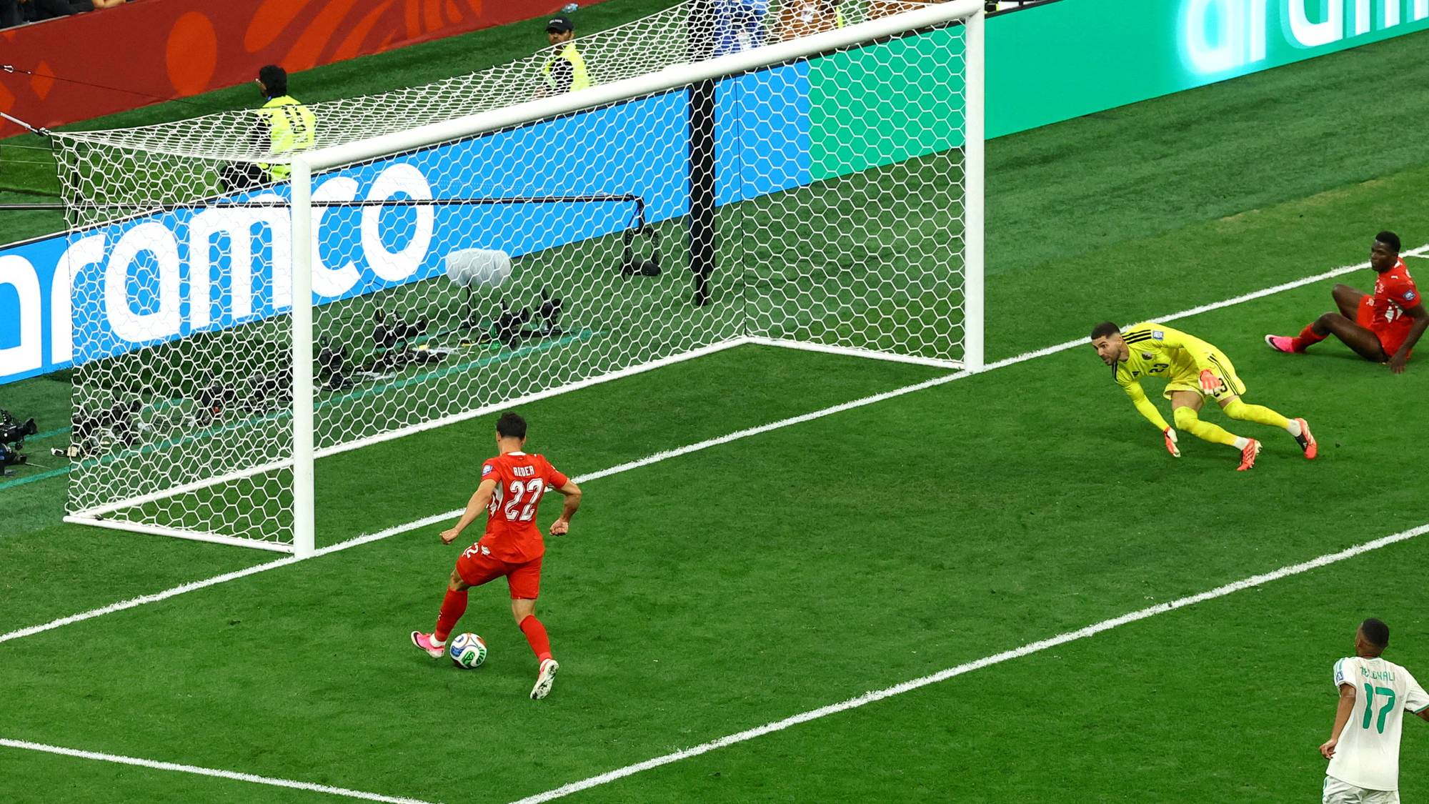 Fotballspiller i rød drakt skyter mot et mål der keeperen kaster seg. Medspiller sitter i gresset. (Bildebeskrivelsen er laget av en KI-tjeneste)