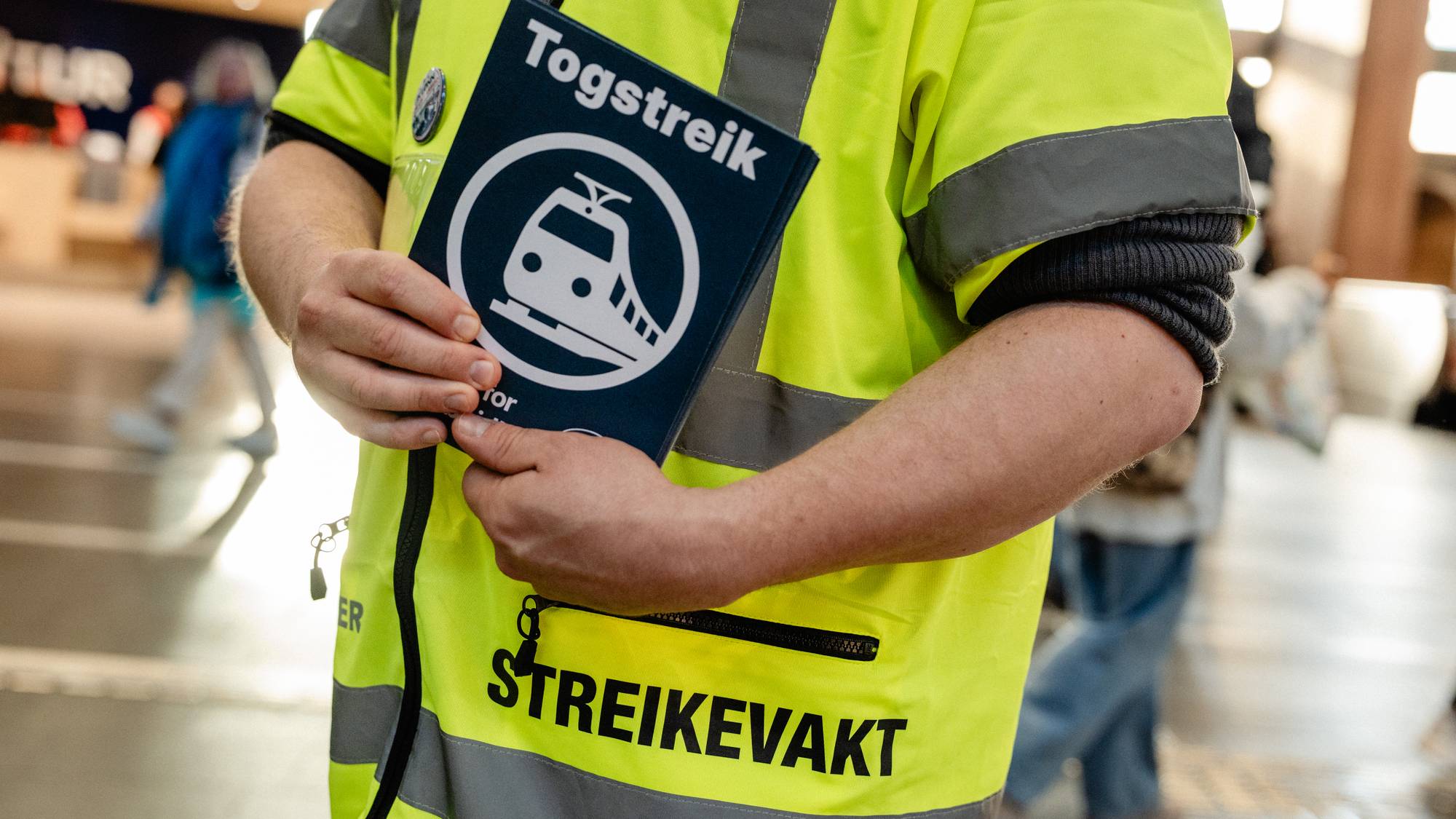 Person i gul refleksvest merket "STREIKEVAKT" holder et skilt med teksten "Togstreik". (Bildebeskrivelsen er laget av en KI-tjeneste)