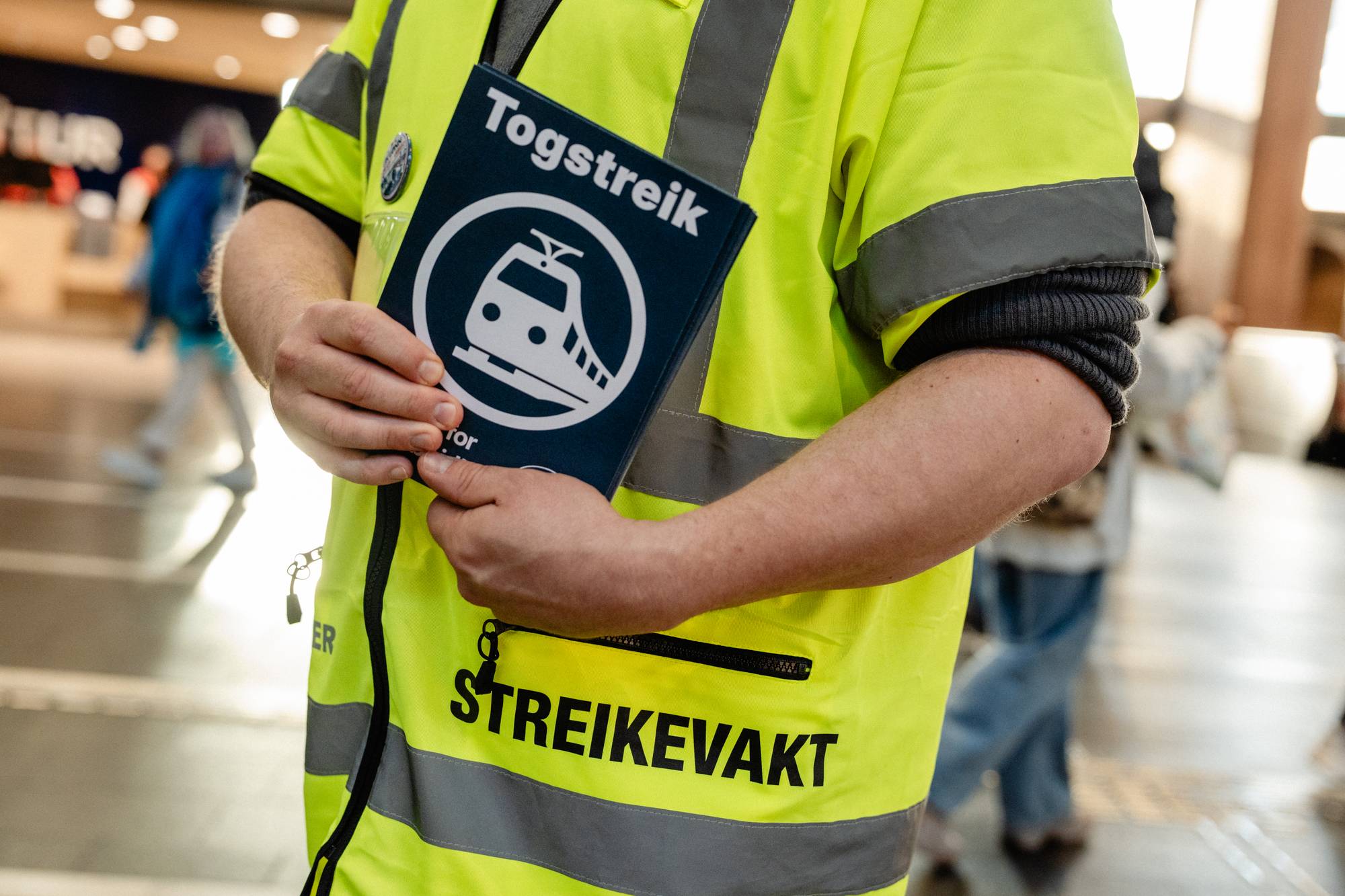 Person i gul refleksvest merket "STREIKEVAKT" holder et skilt med teksten "Togstreik". (Bildebeskrivelsen er laget av en KI-tjeneste)