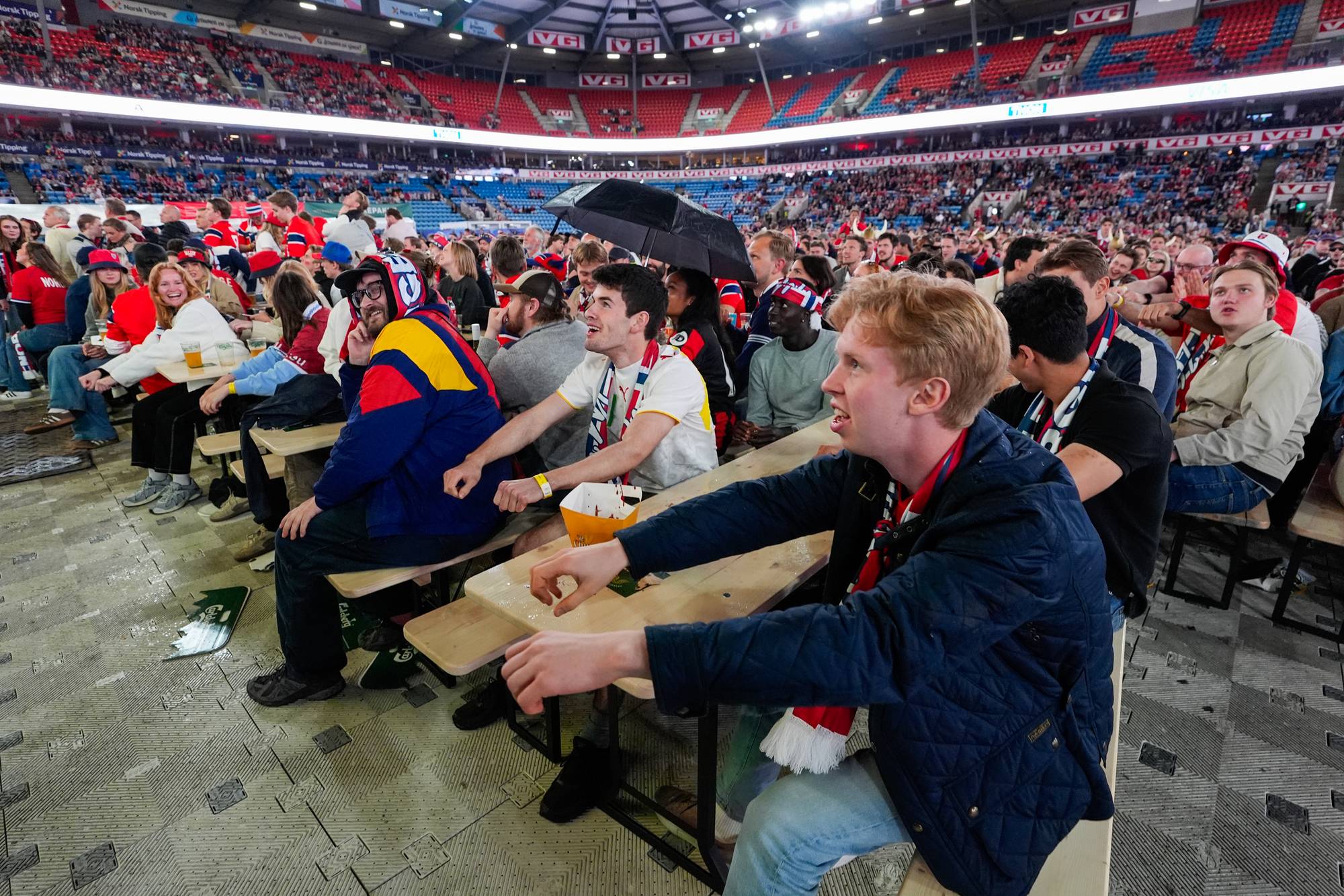 Publikum sitter ved benkerader på en stor arena. En person holder en sort paraply innendørs. (Bildebeskrivelsen er laget av en KI-tjeneste)