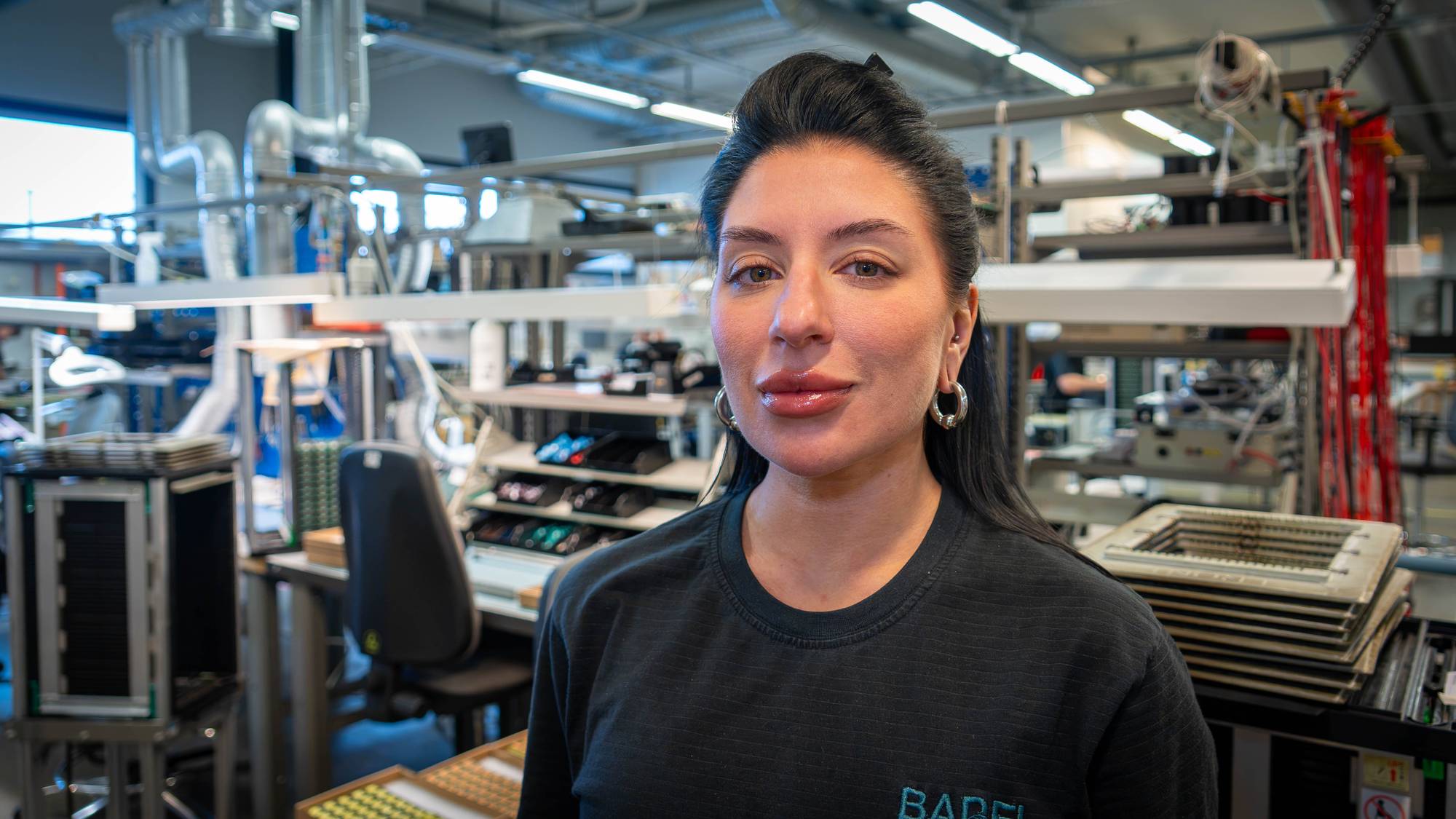 Kvinne i svart genser står i et teknisk verksted eller fabrikk. (Bildebeskrivelsen er laget av en KI-tjeneste)