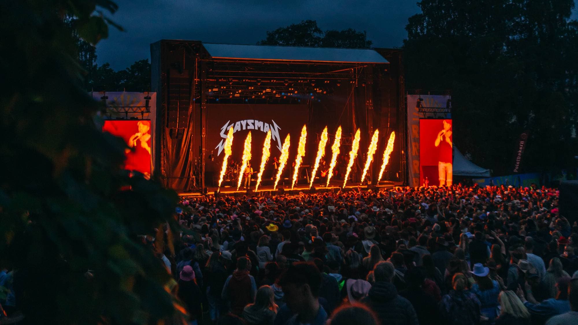 Festival publikum ser på en scene med en artist og flammeeffekter. Scenen har en bakgrunn med tekst og skjermer. Om natten, mens folkemengden er samlet for en konsert, er det flammer som skyter opp fra scenen. (Bildebeskrivelsen er laget av en KI-tjeneste)