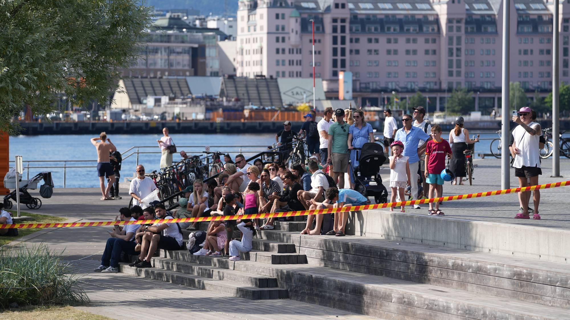 Folk sitter på en amfitrapp ved Oslofjorden avstengt av markeringsbånd. Bygninger i bakgrunnen. (Bildebeskrivelsen er laget av en KI-tjeneste)