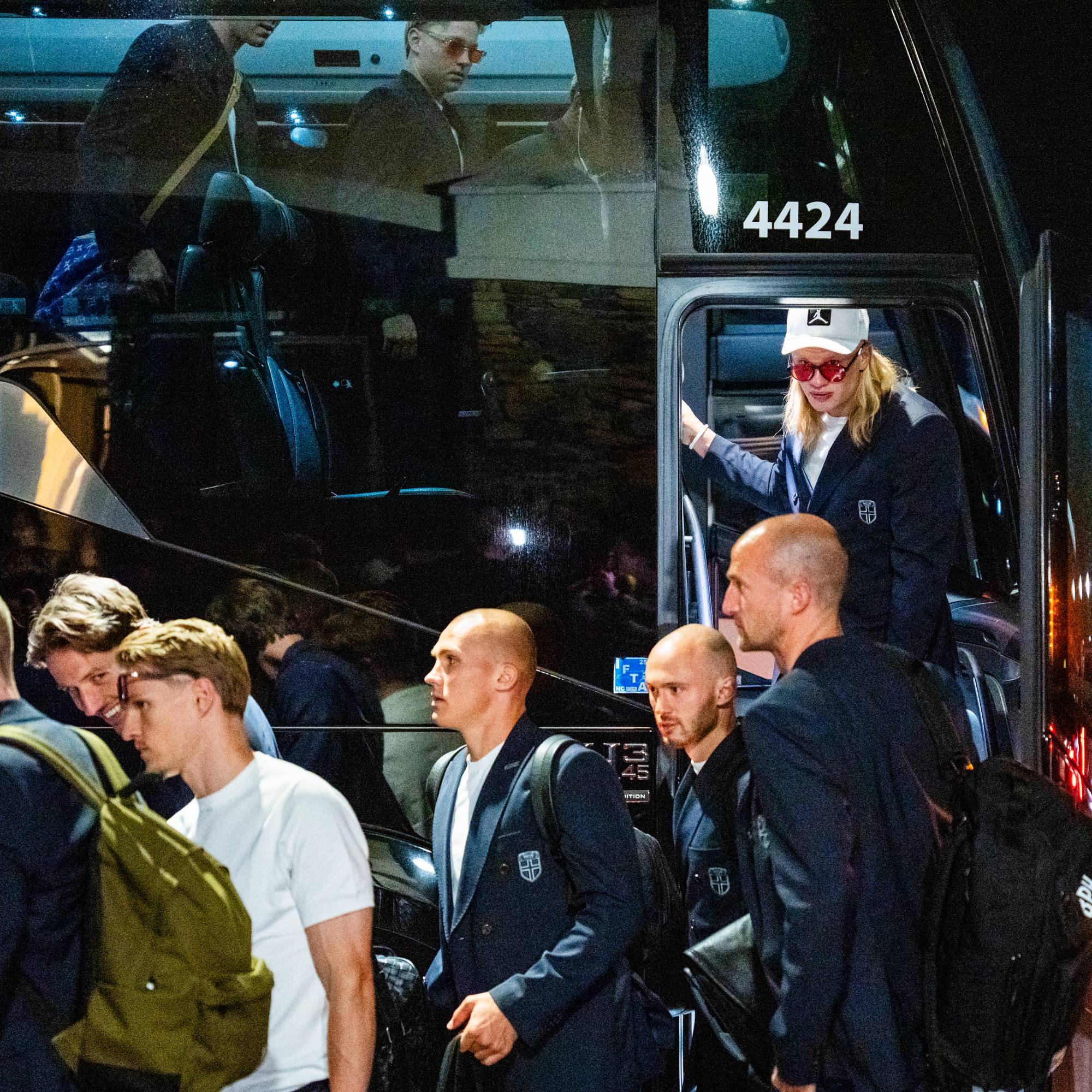 Personer stiger av en buss om natten. En person med hettegenser, caps og solbriller går ned trappen fra bussen. Flere menn i mørke dresser og jakker står foran bussen. (Bildebeskrivelsen er laget av en KI-tjeneste)