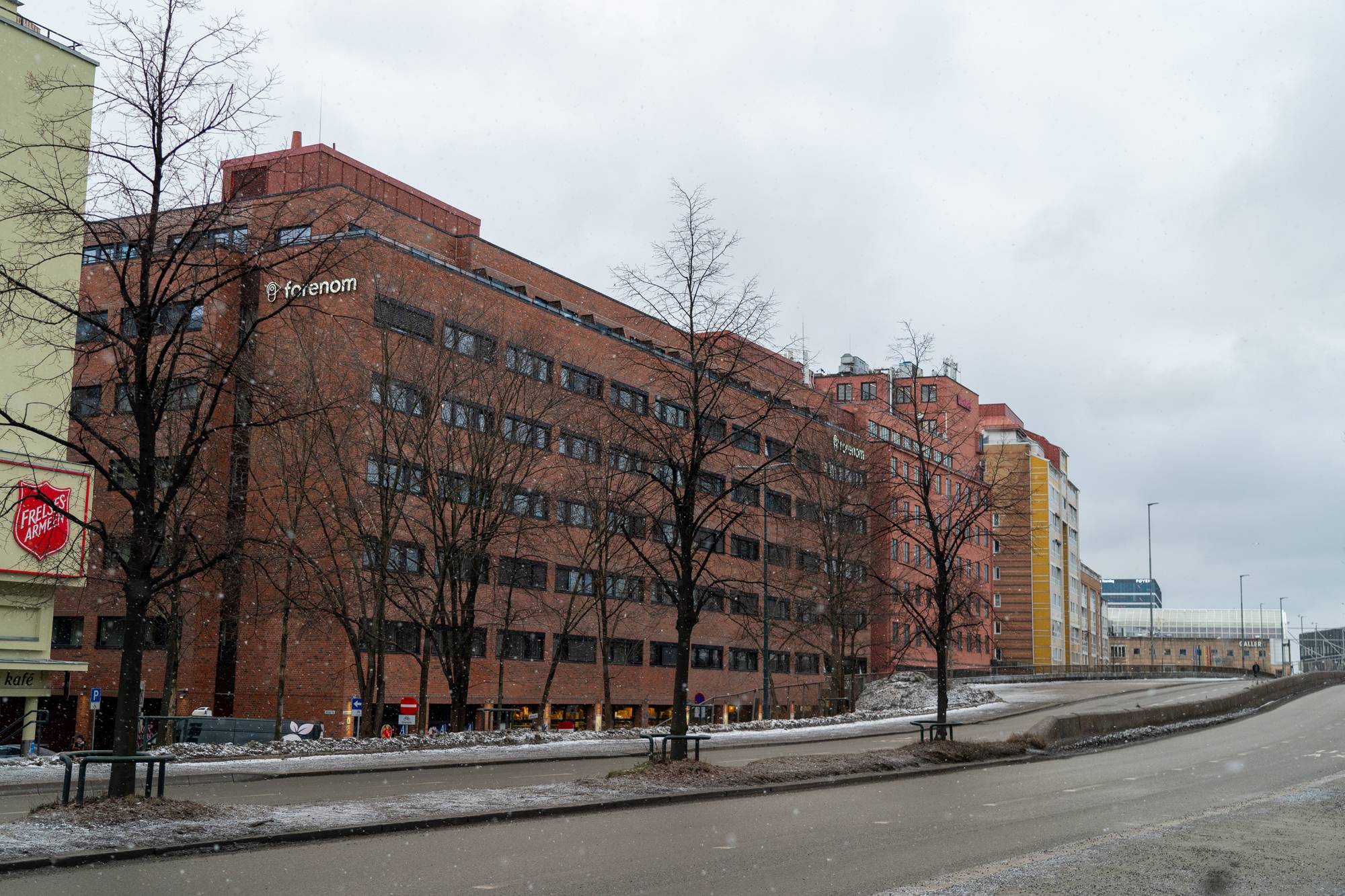 Bildet viser en gate med flere bygninger langs siden. Den mest fremtredende bygningen har en rød mursteinsfasade. Det er snøfall.