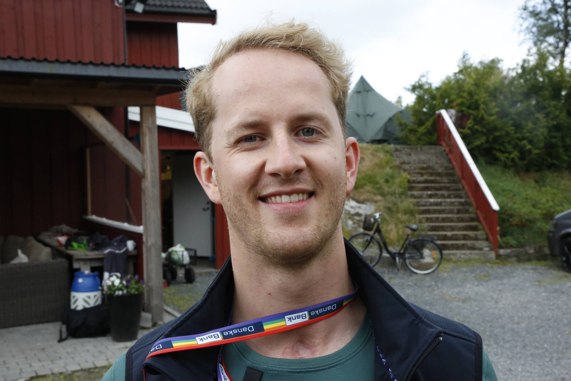 Portrett av en smilende mann med et regnbuefarget bånd rundt halsen utendørs. (Bildebeskrivelsen er laget av en KI-tjeneste)