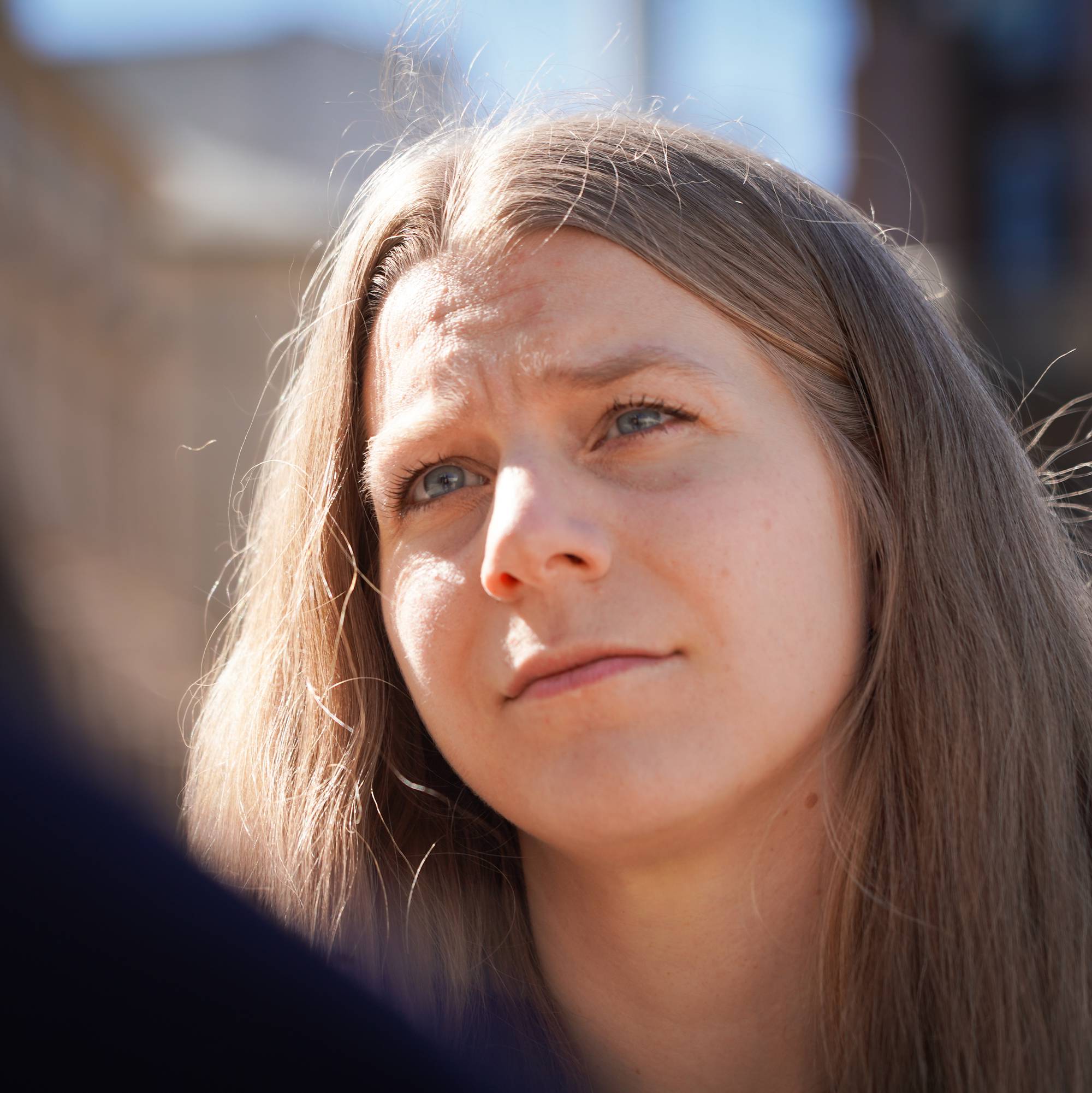 En kvinne med langt, lyst hår ser til siden med bekymret mine. Hun er delvis tildekket av en person foran. I bakgrunnen sees en bygning og en blå himmel. (Bildebeskrivelsen er laget av en KI-tjeneste)