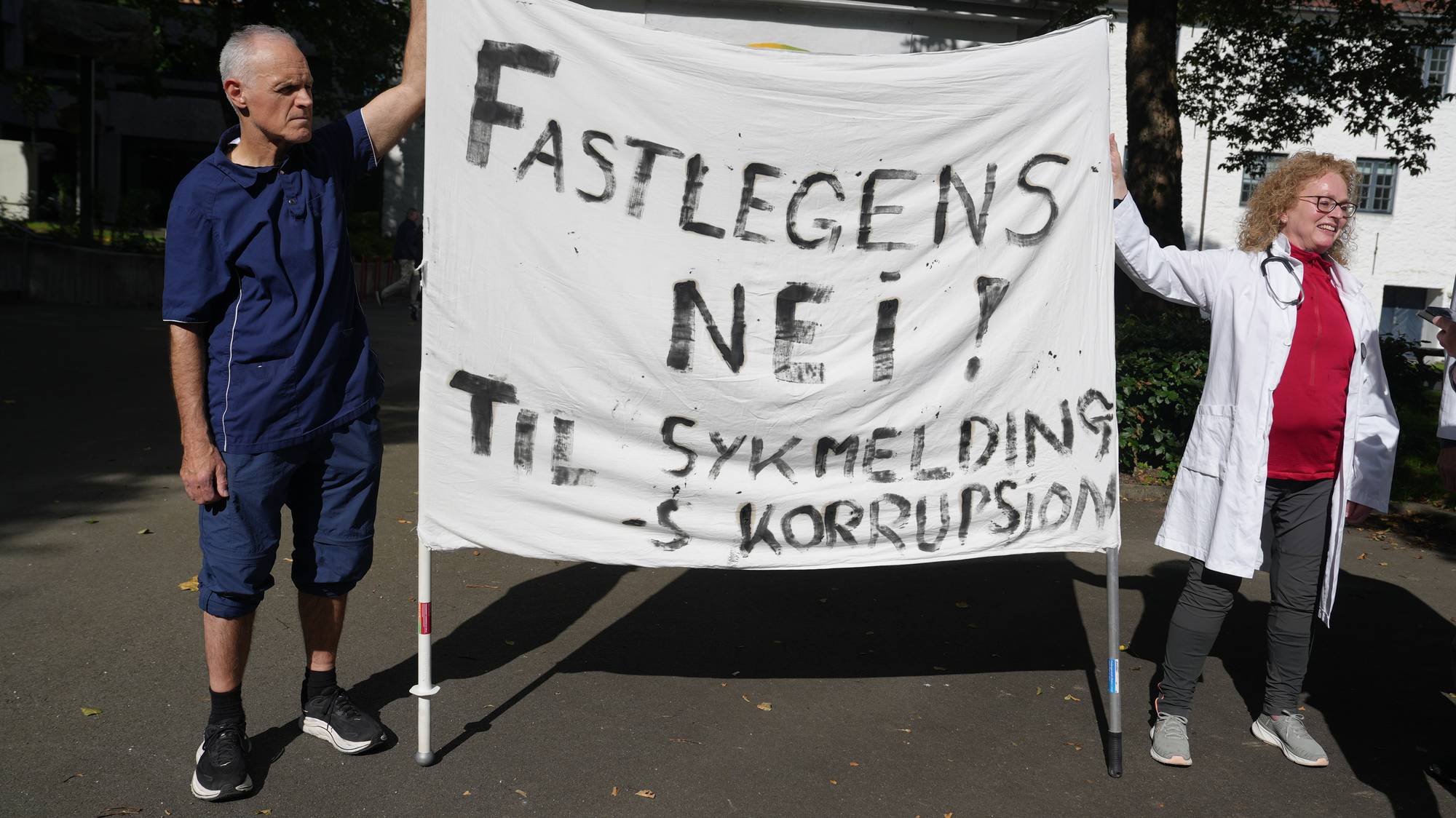 To personer i helseuniform holder en plakat med teksten Fastlegens nei til sykmeldingskorrupsjon. (Bildebeskrivelsen er laget av en KI-tjeneste)