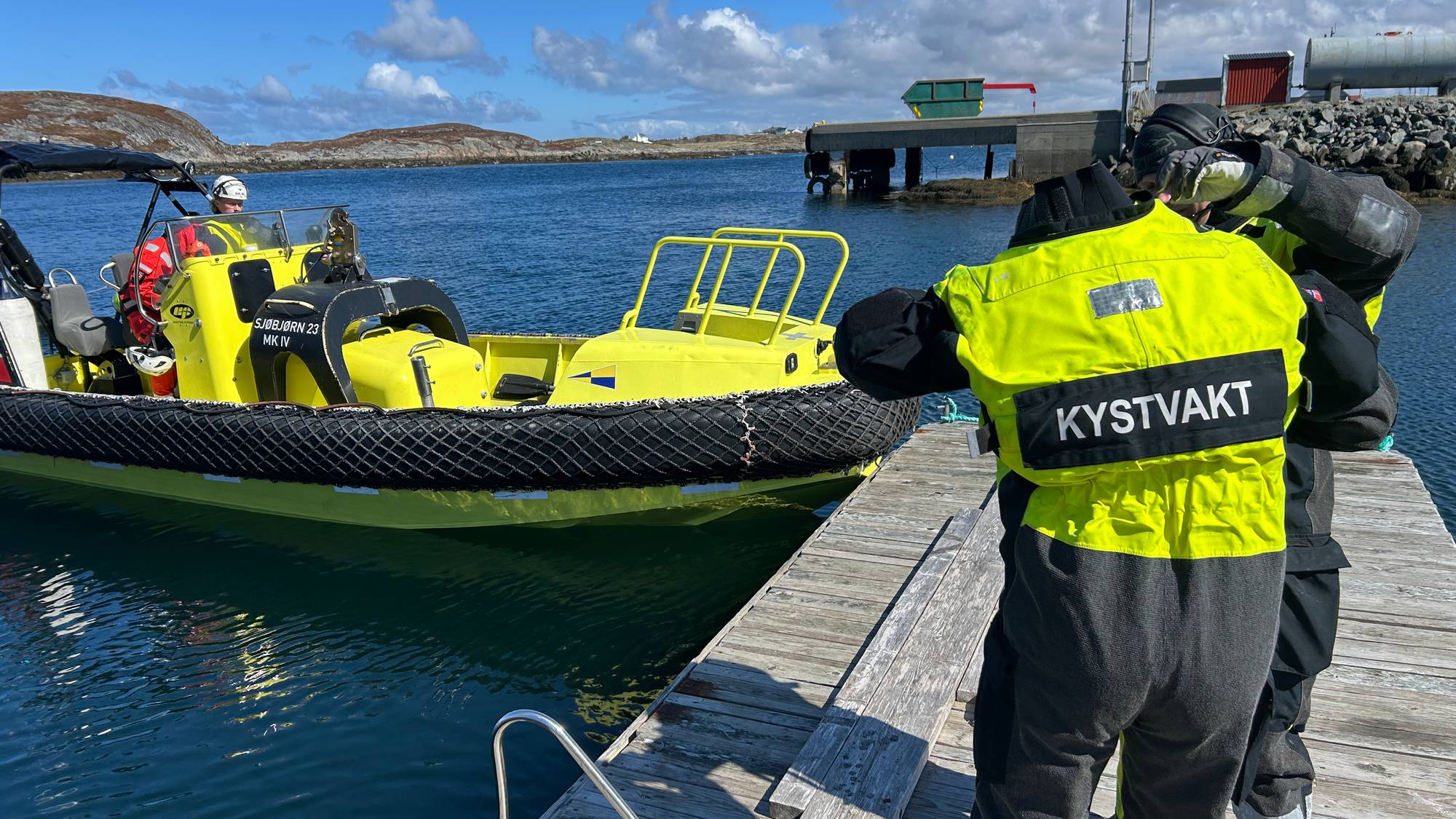 Kystvaktpersonell i gule vester står på et bryggeanlegg. En gul redningsbåt ligger fortøyd ved bryggen. Bak dem er det en kai med industriell infrastruktur. Solen skinner på en klar himmel. (Bildebeskrivelsen er laget av en KI-tjeneste)