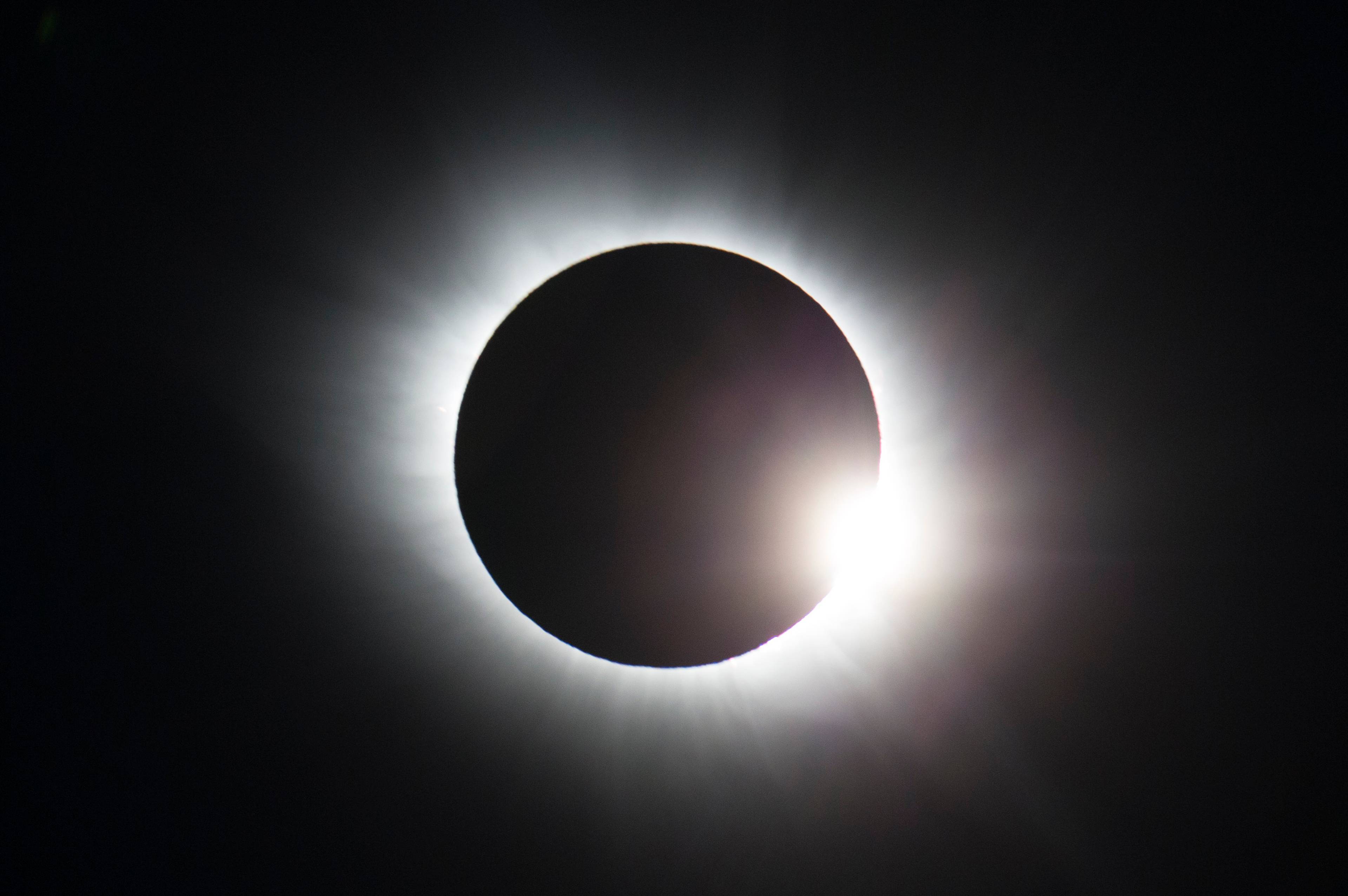 Bildet viser en delvis solformørkelse der månen delvis dekker solen. Lysstråler renner ut rundt månens konturer, noe som skaper en glorie-effekt. Bakgrunnen er svart, noe som fremhever kontrasten mellom solen og månen. Dette astronomiske fenomenet er sett av mange observatører verden over. (Bildebeskrivelsen er laget av en KI-tjeneste)