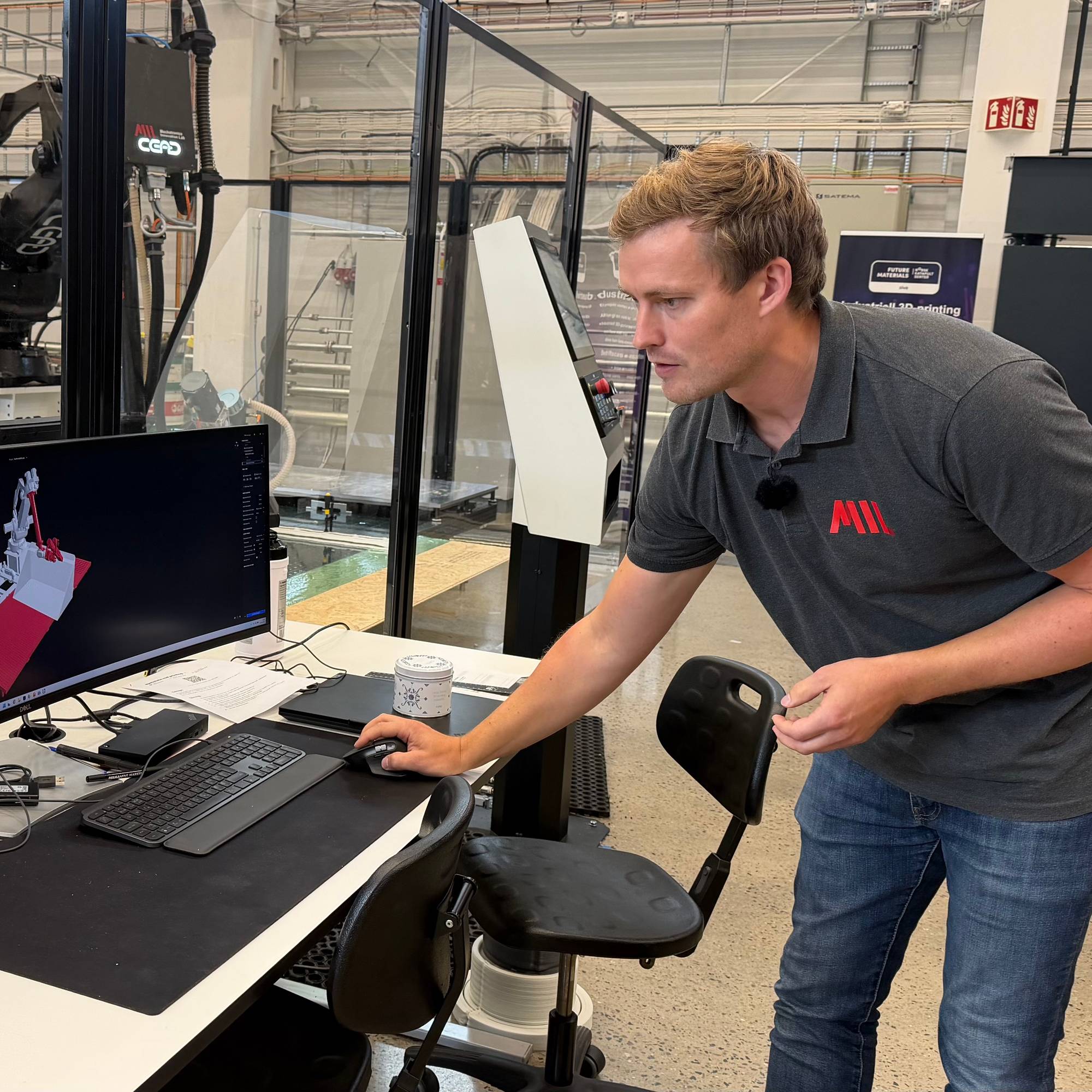 Man i svart genser viser dataskjerm. Er en stor robot/3D printer i bakgrunnen.