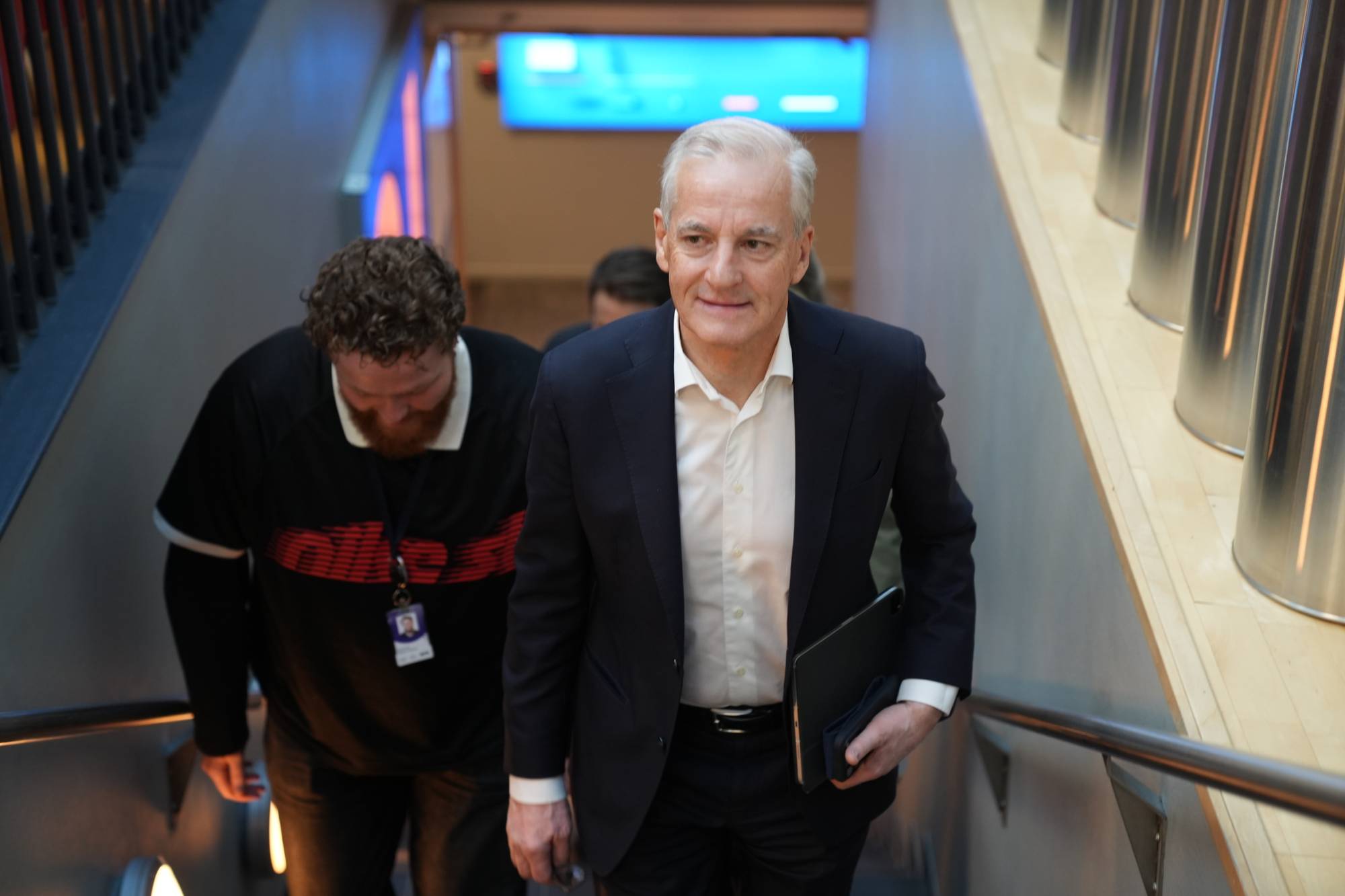 En mann i mørk dress går opp en trapp. Han holder en nettbrett. Bak ham går en annen mann i en svart overdel. De befinner seg innendørs. (Bildebeskrivelsen er laget av en KI-tjeneste)