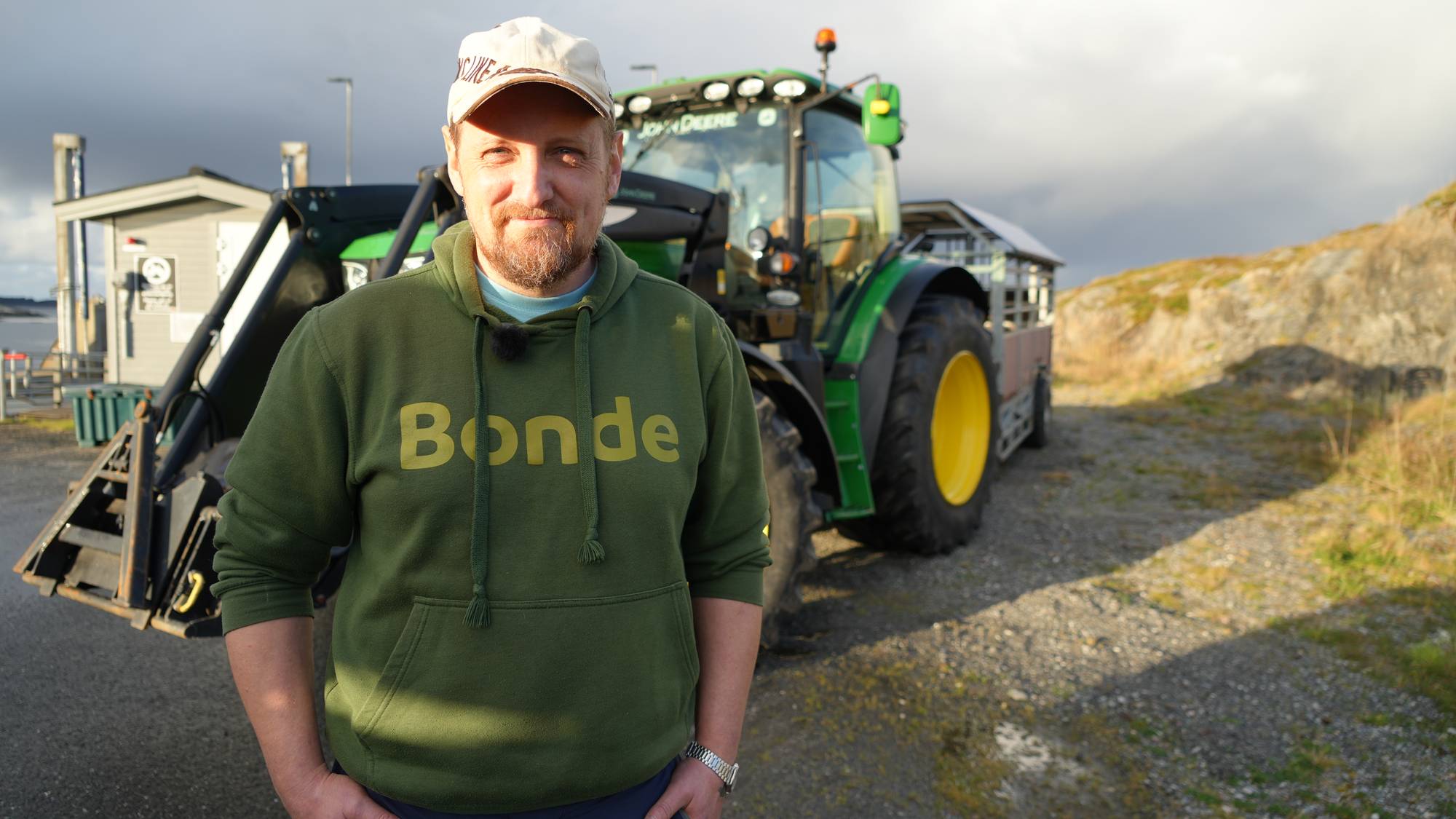 En bonde i grønn genser står foran en traktor ved havet. (Bildebeskrivelsen er laget av en KI-tjeneste)