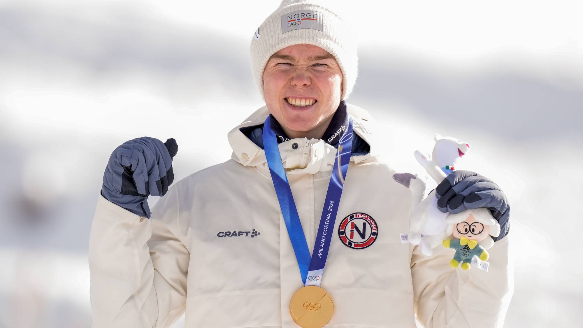 En utøver poserer stolt med en gullmedalje rundt halsen og holder en bamse i den ene hånden. Utøveren har på seg en hvit jakke med norsk flagg og er iført hansker. Bakgrunnen viser snødekte fjell i klart vær. Medaljen er fra et internasjonalt mesterskap. (Bildebeskrivelsen er laget av en KI-tjeneste)