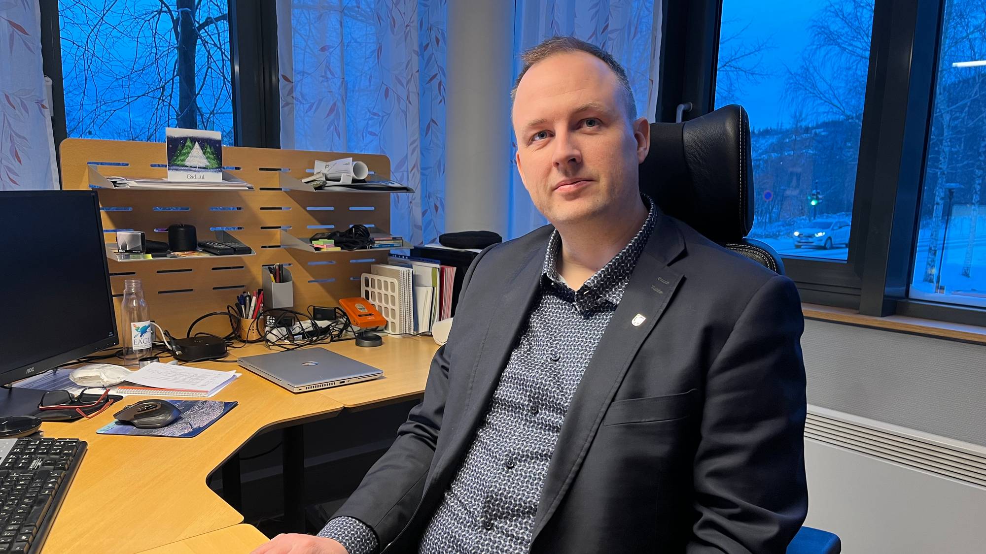 En mann sitter ved et kontorbord med en datamaskin og diverse kontorrekvisita. Utenfor vinduet er det snø og et kjøretøy kjører forbi. Mannen har på seg dressjakke og skjorte. (Bildebeskrivelsen er laget av en KI-tjeneste)