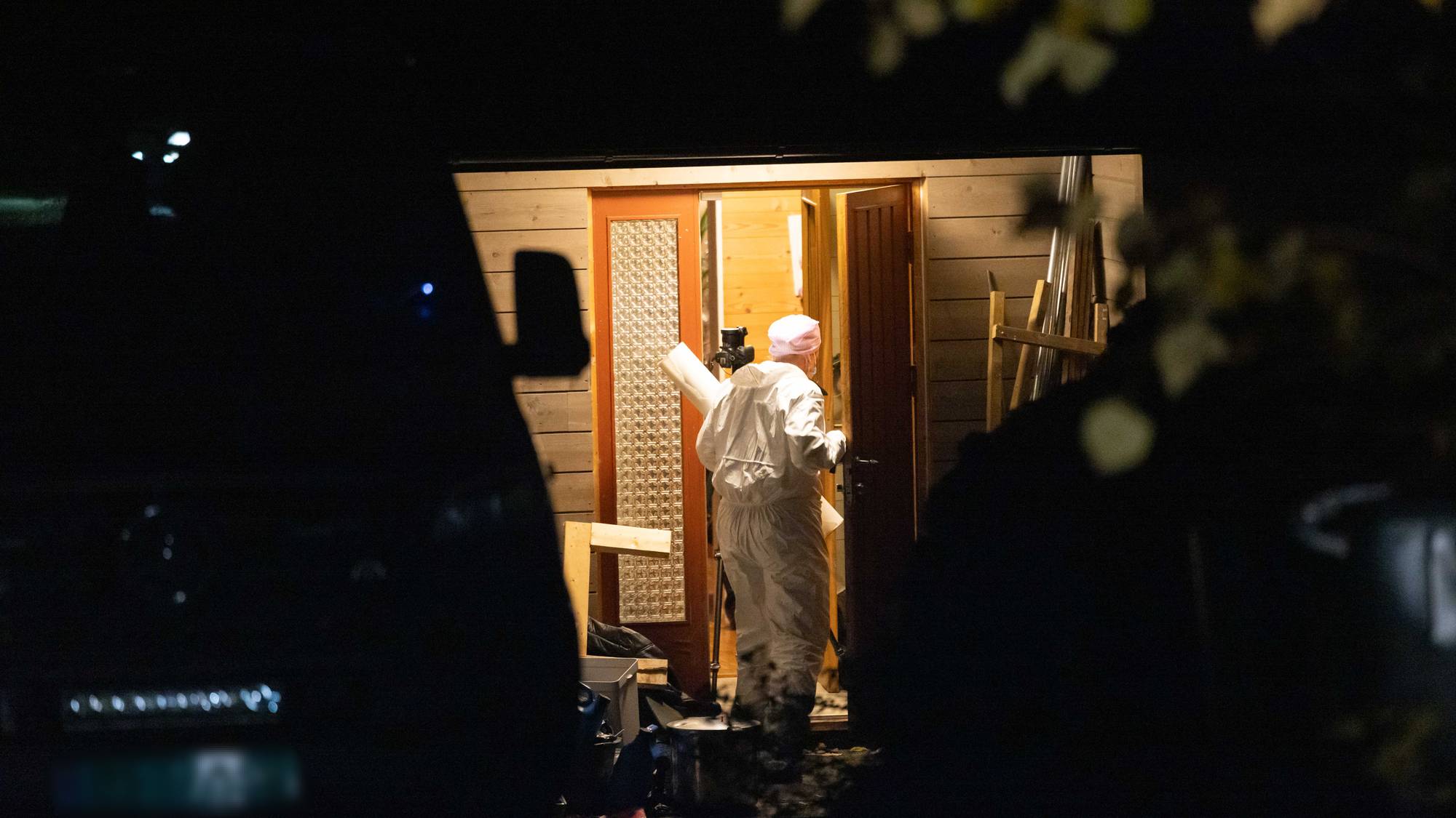 En politteknikeri hvit overtrekksdress er på vei inn hoveddøren i et hus. Det er lyst over inngangspartiet, men mørkt i omgivelsene. 