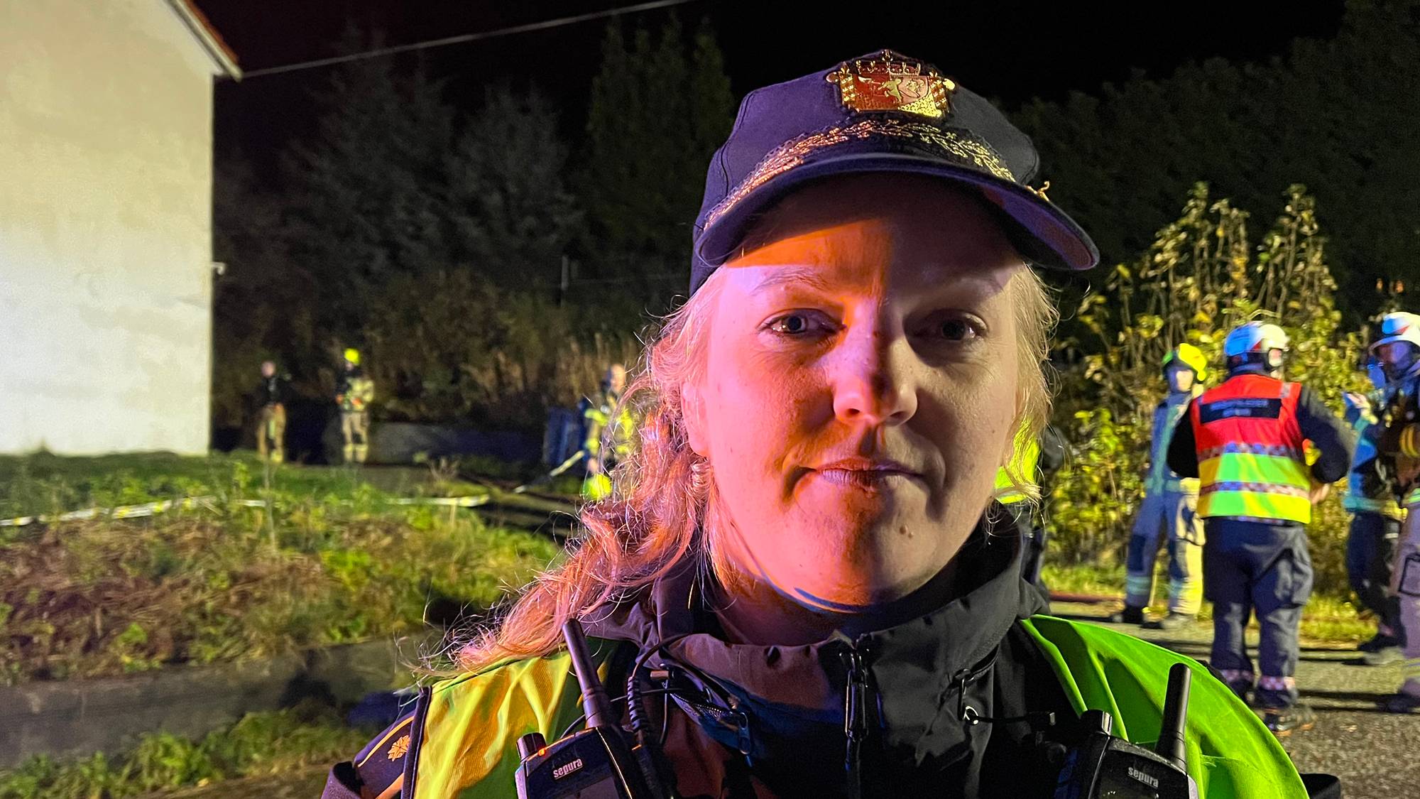 En kvinnelig politibetjent i uniform med samband på brystet på et åsted om natten. (Bildebeskrivelsen er laget av en KI-tjeneste)