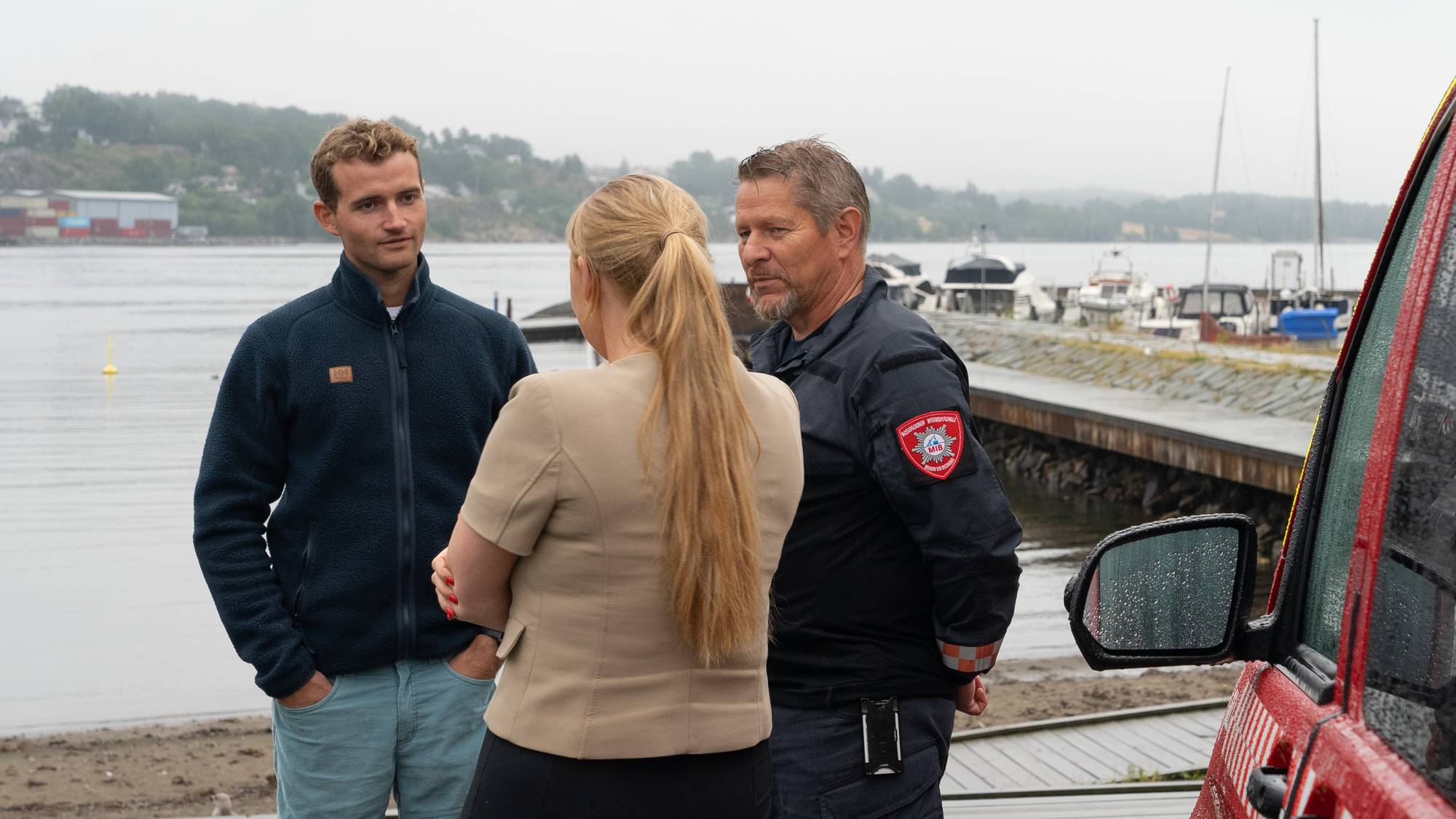 En brannmann snakker med to personer ved en brygge med en brannbil i forgrunnen. (Bildebeskrivelsen er laget av en KI-tjeneste)