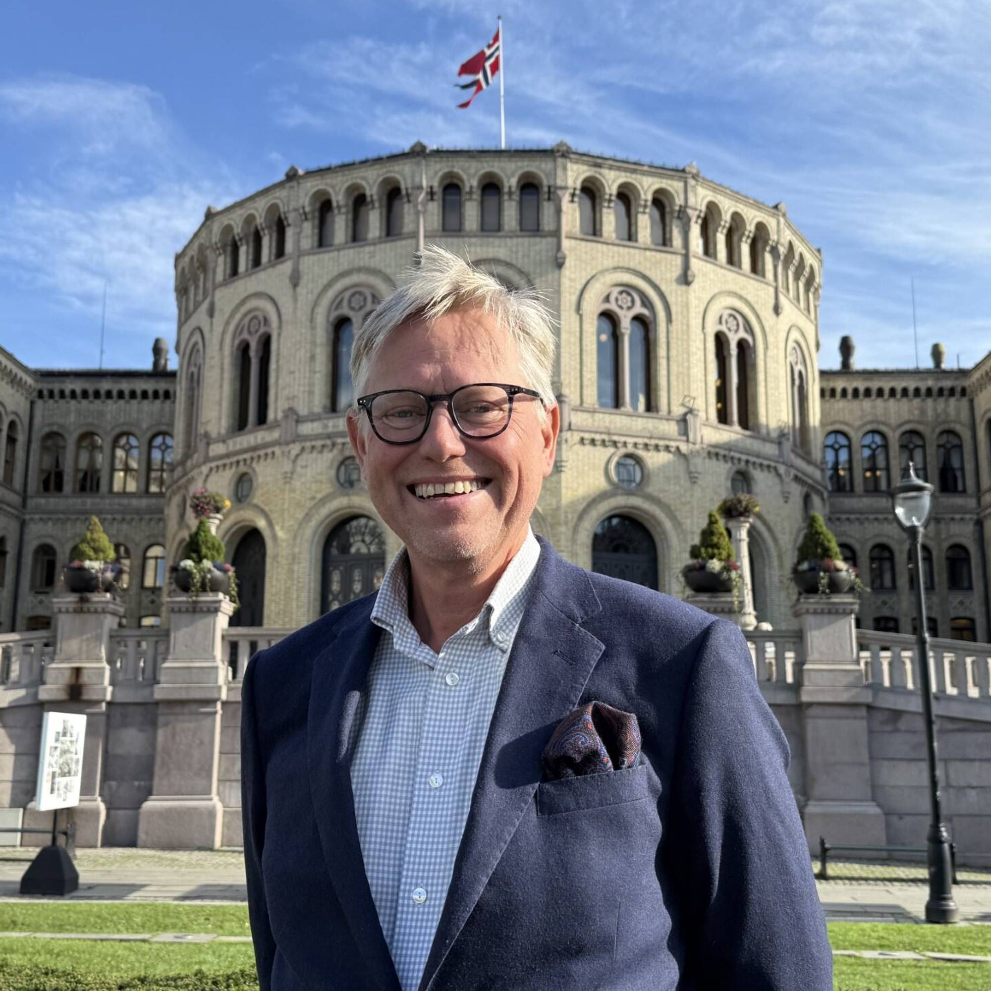 Bildet viser en mann med briller som smiler. Han har på seg dress ogs står foran Stortinget.