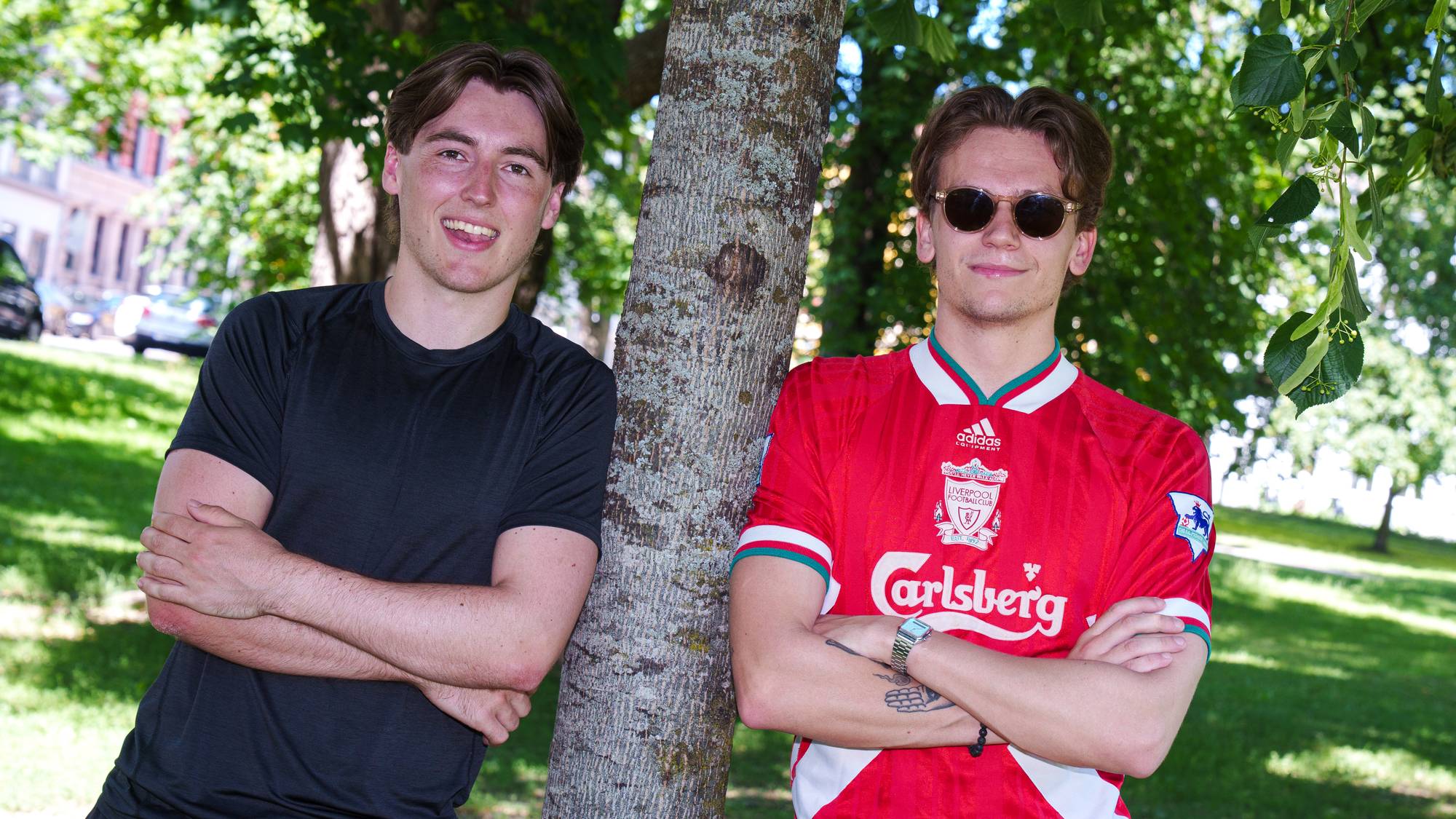 Portrett av to unge menn som står inntil en trestamme. En av dem smiler bredt. Den andre smiler lurt og har på seg solbriller og en fotballdrakt.
