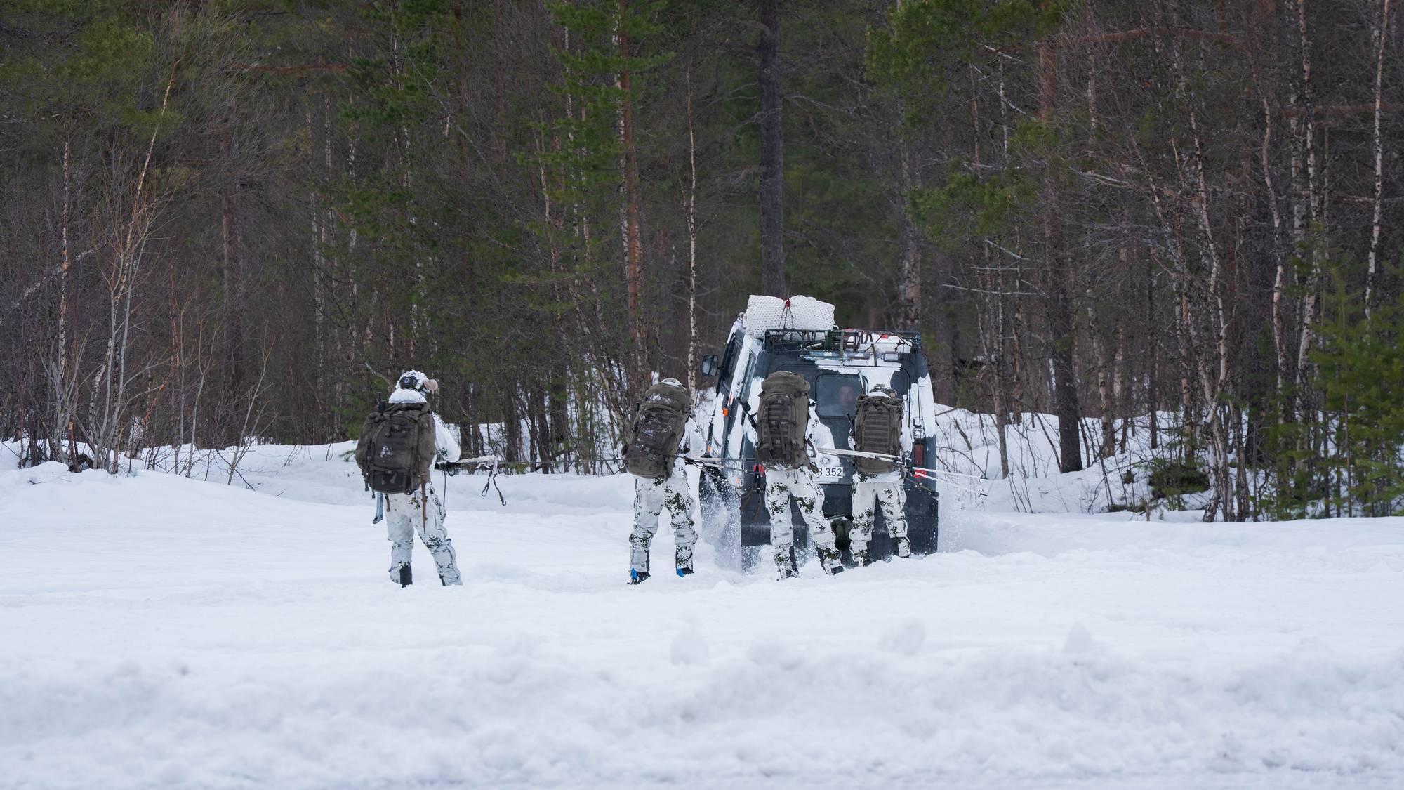 Soldater i kamuflasjedrakter med store ryggsekker nærmer seg et militært kjøretøy i snødekt terreng. De er ute i en skogkledd lysning. (Bildebeskrivelsen er laget av en KI-tjeneste)