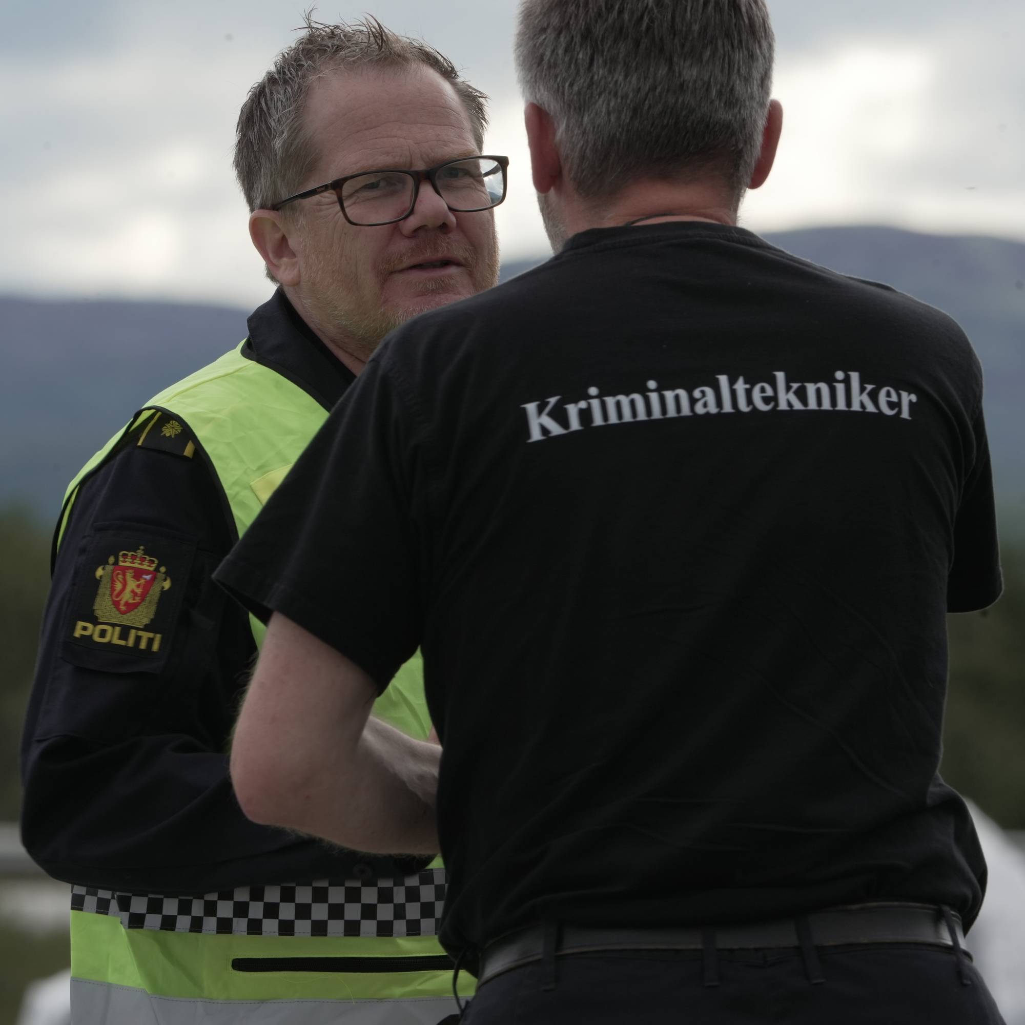 En politibetjent i refleksvest snakker med en kriminaltekniker utendørs. 