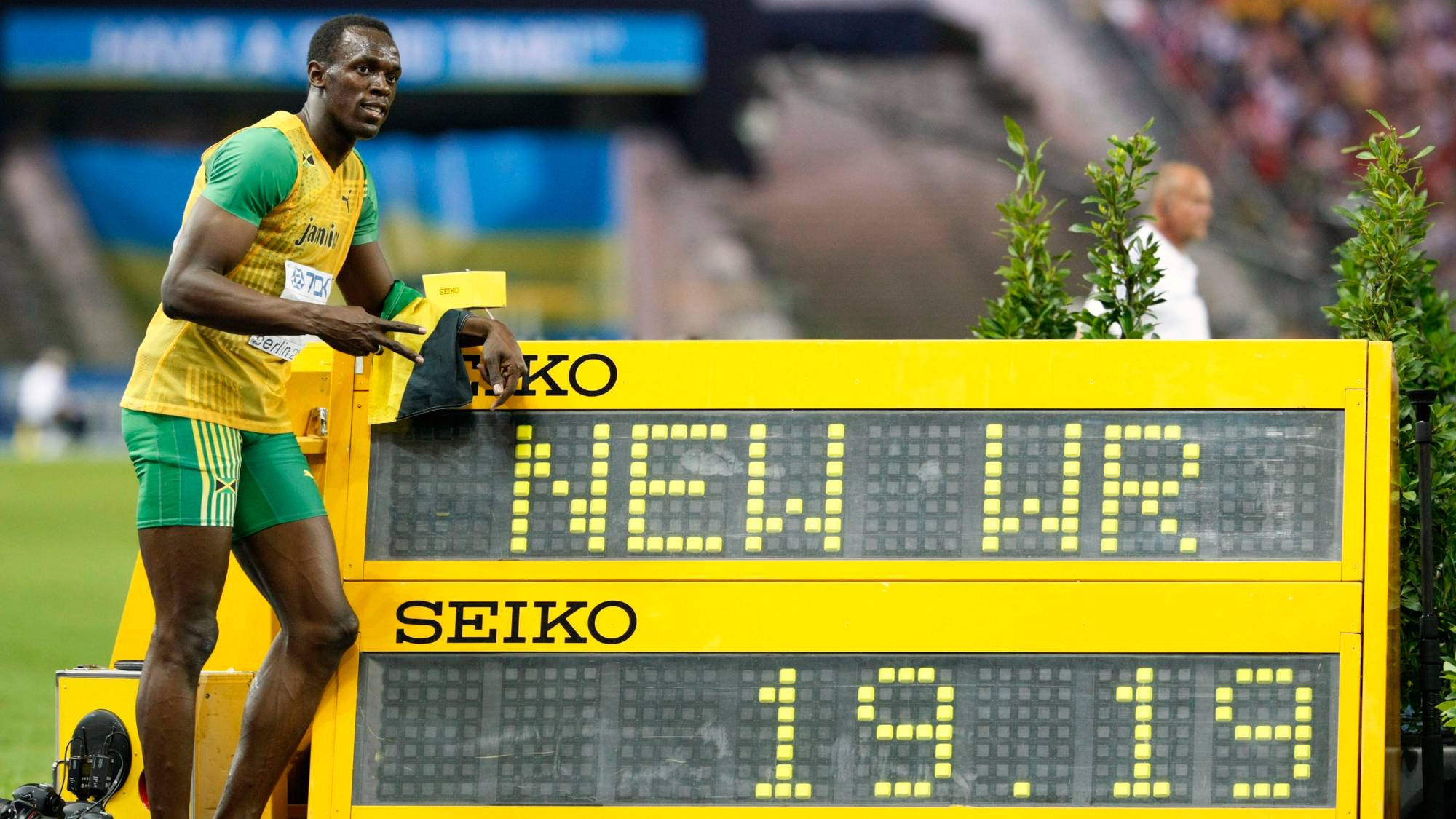 Usain Bolt står ved siden av et resultatbrett som viser 19.19. Brettet viser også tekst. En mann i hvit skjorte går forbi i bakgrunnen. Det er en idrettsbane og tribuner i bakgrunnen. (Bildebeskrivelsen er laget av en KI-tjeneste)