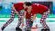 LYLE STAFFORD / Reuters / NTB Tre personer deltar i curling på en isflate under et mesterskap. De bærer røde t-skjorter og bukser med et sjakkrutet mønster i rødt, hvitt og blått. En av dem er i ferd med å løfte en curlingstein, mens de andre forbereder seg til å gi instruksjoner. Området rundt dem er innredet for curling-aktiviteter, med en tydelig isbane og tilskuere i bakgrunnen. (Bildebeskrivelsen er laget av en KI-tjeneste)