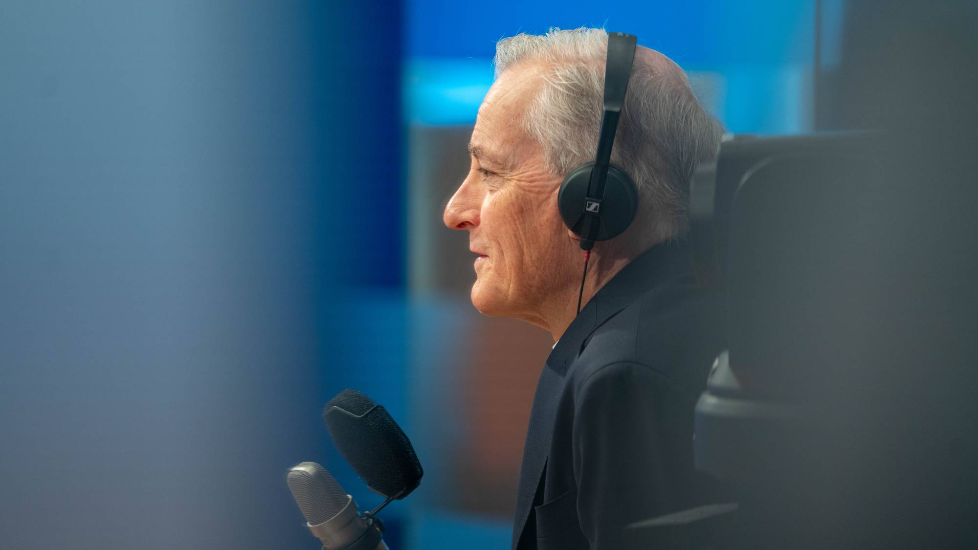 En eldre mann med hodetelefoner snakker inn i en mikrofon. Bakgrunnen er blå og uskarpt. Dette kan være en radio- eller podkast-innspilling. (Bildebeskrivelsen er laget av en KI-tjeneste)