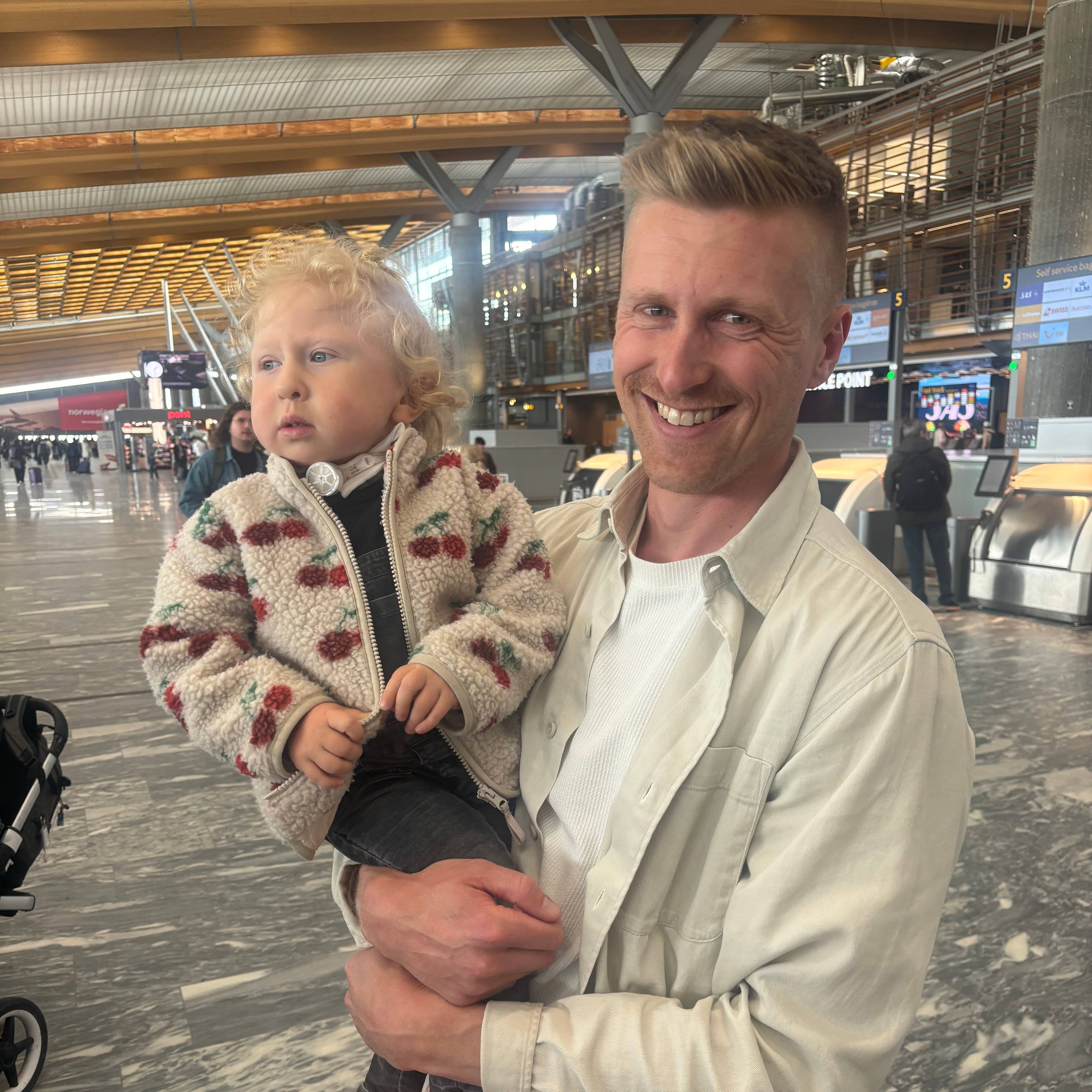 En mann holder et lite barn på en flyplass. Mannen smiler mens barnet ser til siden.