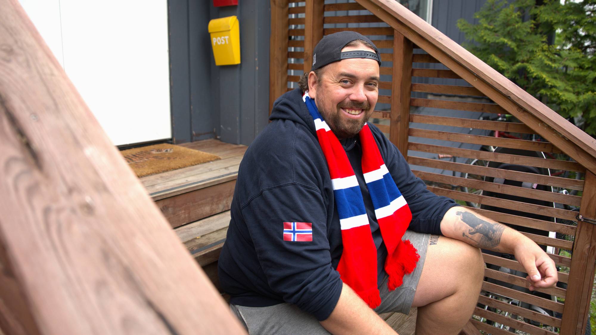 Mann sitter på trappa foran en hvit dør med hettegenser, caps og et rødt, hvitt og blått supporterskjerf og smiler.
