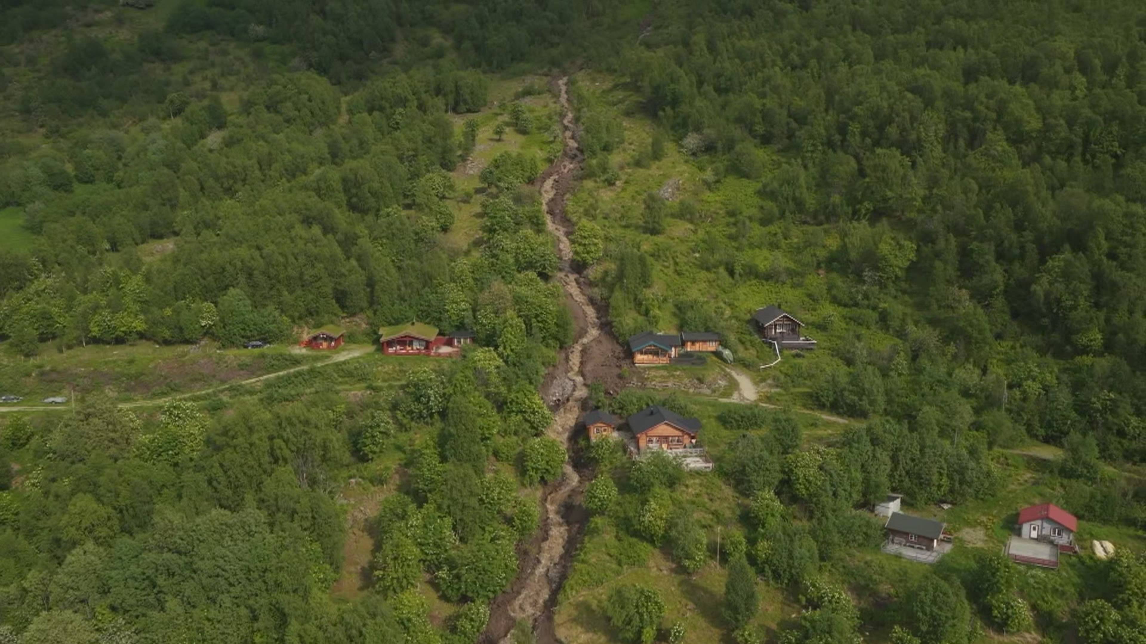 En flom har rennet nedover en skråning med trær og hus. Den mudrete vannføringen skiller seg ut fra det grønne landskapet. Flere hus, noen røde og andre i tre, er spredt langs skråningen. (Bildebeskrivelsen er laget av en KI-tjeneste)