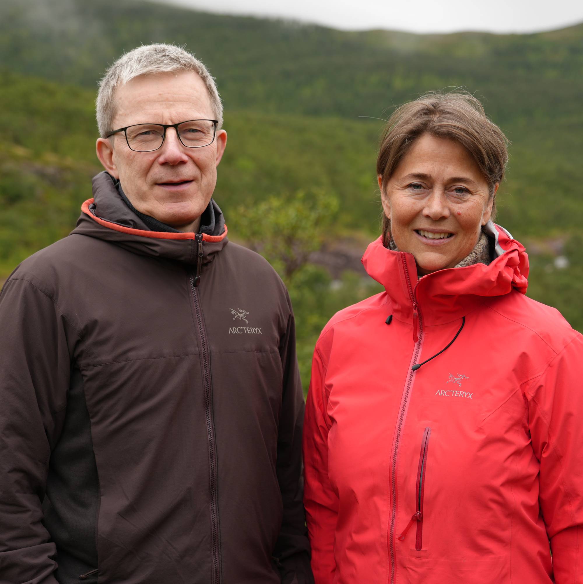 En mann og en kvinne står på en eng med trær og åser i bakgrunnen (Bildebeskrivelsen er laget av en KI-tjeneste)