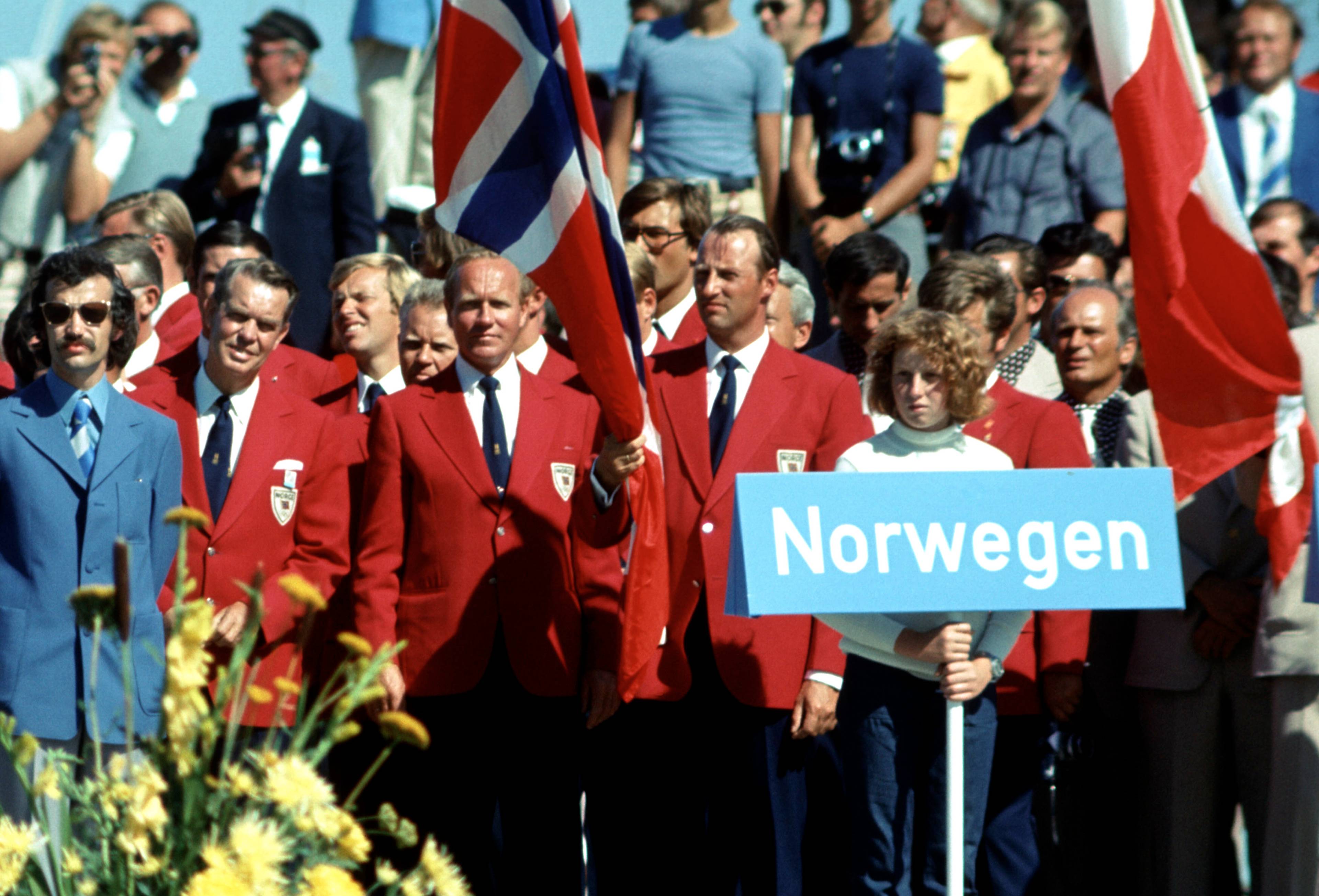 Norsk delegasjon med flagg og skilt under OL i Kiel i 1972. Personer i blazer står på en tribune. (Bildebeskrivelsen er laget av en KI-tjeneste)