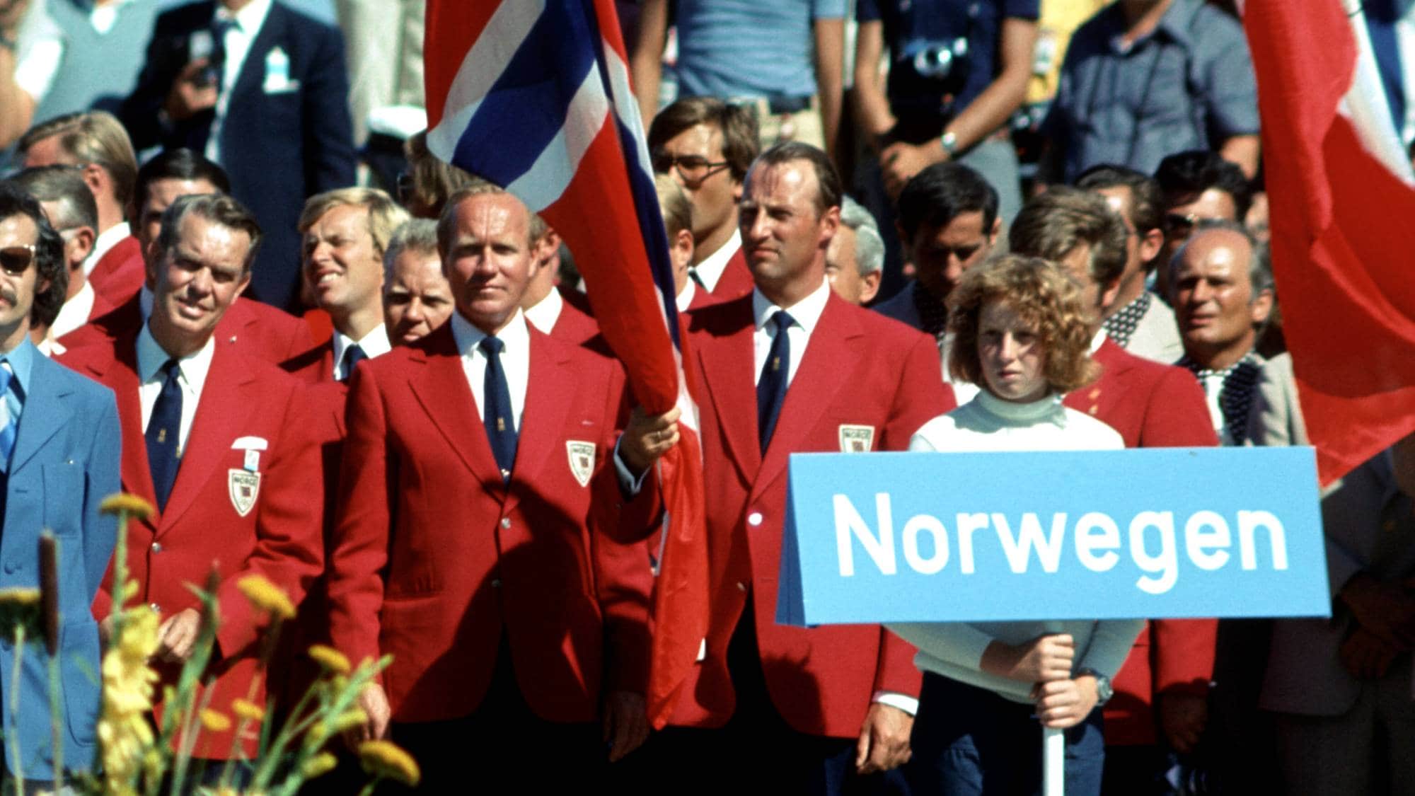 Norsk delegasjon med flagg og skilt under OL i Kiel i 1972. Personer i blazer står på en tribune. (Bildebeskrivelsen er laget av en KI-tjeneste)