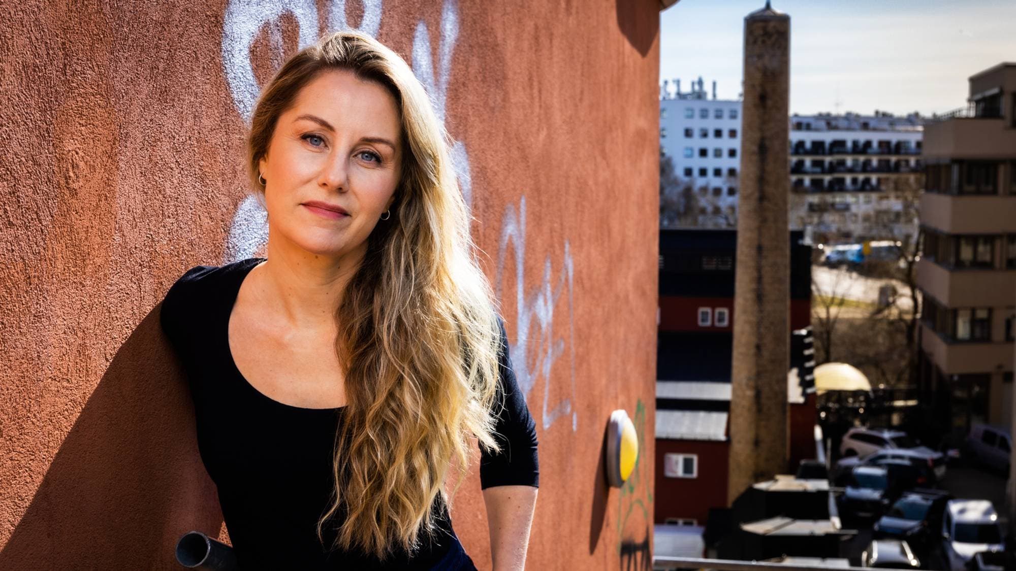 Kaja Melsom i svart genser foran en rustbrun husvegg med bygate i bakgrunnen.