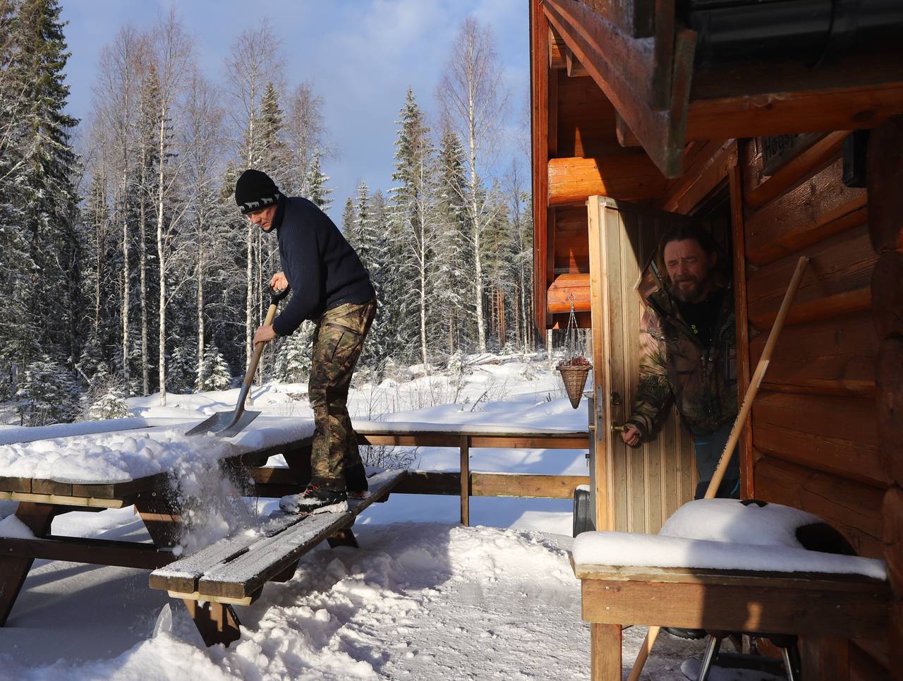 Jan måker snø vekk fra et piknikbord utenfor hytta mens Helge åpner døren og blir blendet av sollyset. 
