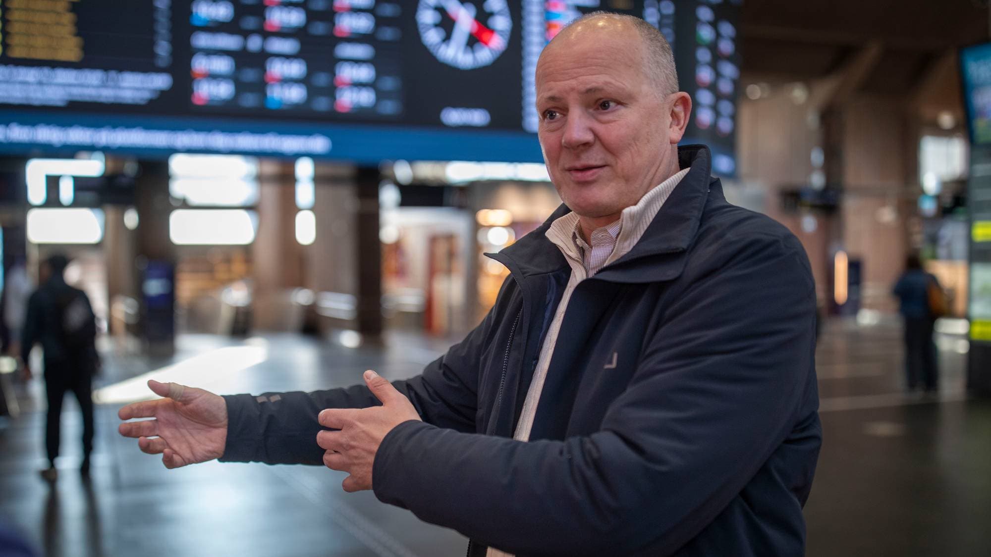 En mann snakker og gestikulerer foran en avgangstavle på en togstasjon. (Bildebeskrivelsen er laget av en KI-tjeneste)