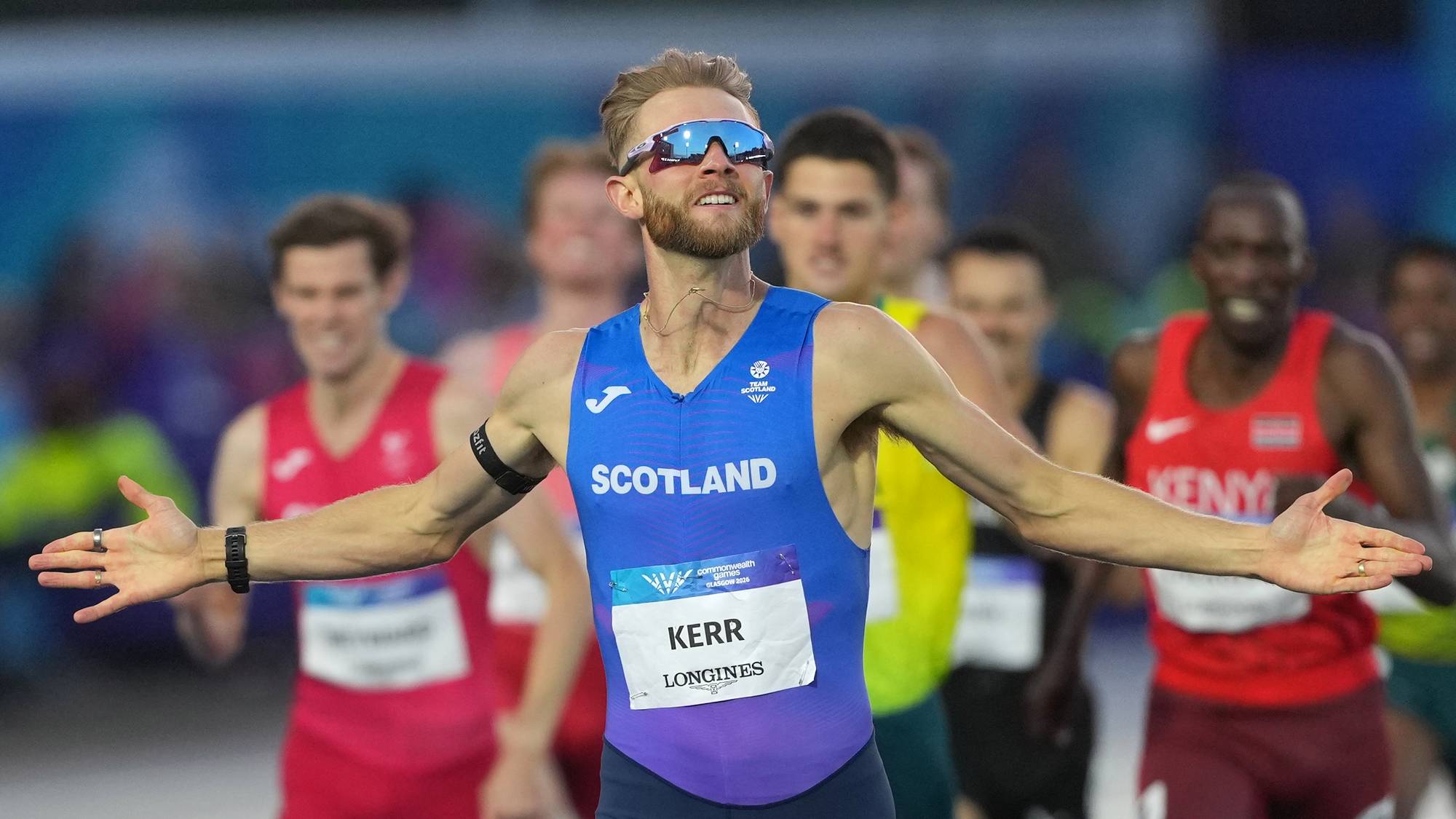 Josh Kerr jubler med utslåtte armer etter et løp på en friidrettsbane med andre løpere bak. (Bildebeskrivelsen er laget av en KI-tjeneste)