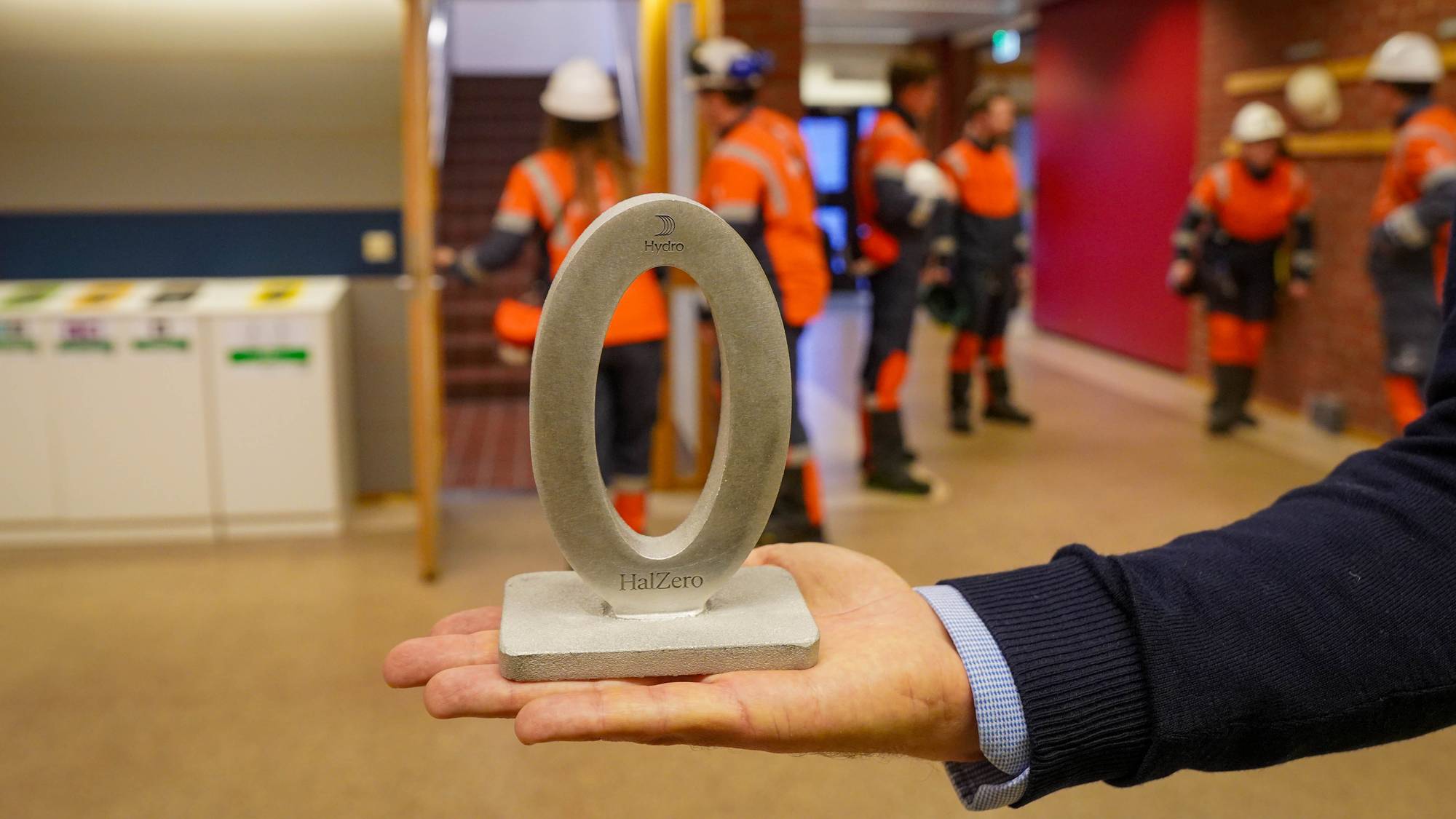 En person holder en HalZero-figur i metall. I bakgrunnen står flere personer i verneutstyr. (Bildebeskrivelsen er laget av en KI-tjeneste)
