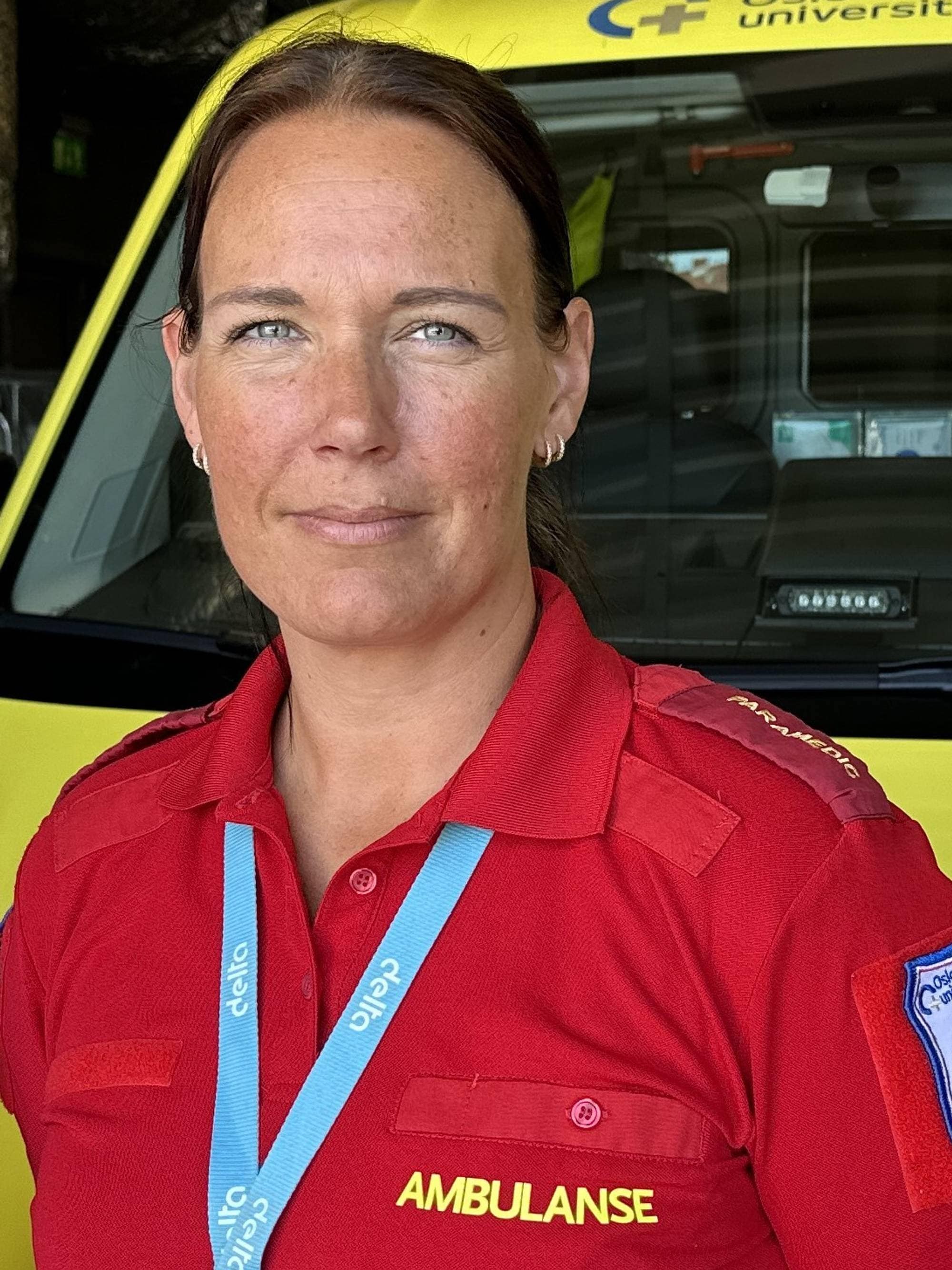 En ambulanseservicemedarbeider står foran en gult ambulansekjøretøy. Hun har på seg en rød uniform med påskriften "AMBULANSE". I bakgrunnen kan deler av ambulansen sees, inkludert seter og utstyr. Det er tydelig at bildet er tatt på en ambulansestasjon. (Bildebeskrivelsen er laget av en KI-tjeneste)