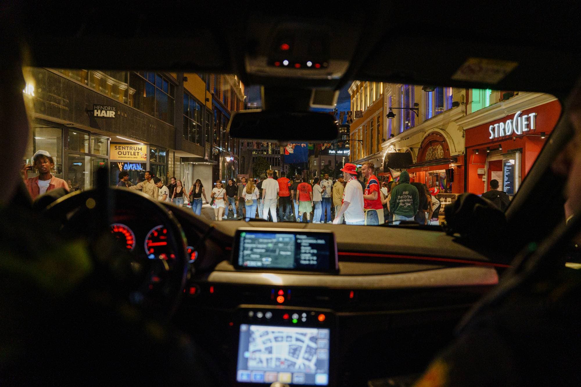 Utsikt fra bilens forsete mot en travel gate med fotgjengere på kveldstid. (Bildebeskrivelsen er laget av en KI-tjeneste)