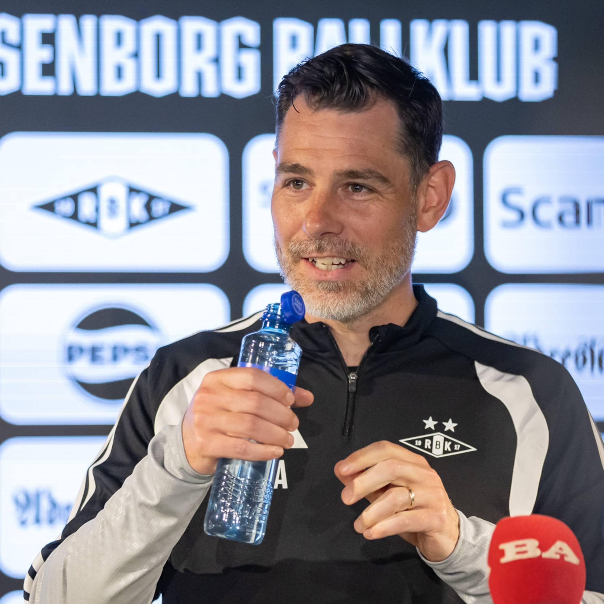 RBK-trener Freyr Alexandersson er på en pressekonferanse. Han er i ferd med å drikke fra en flaske vann. Han smiler