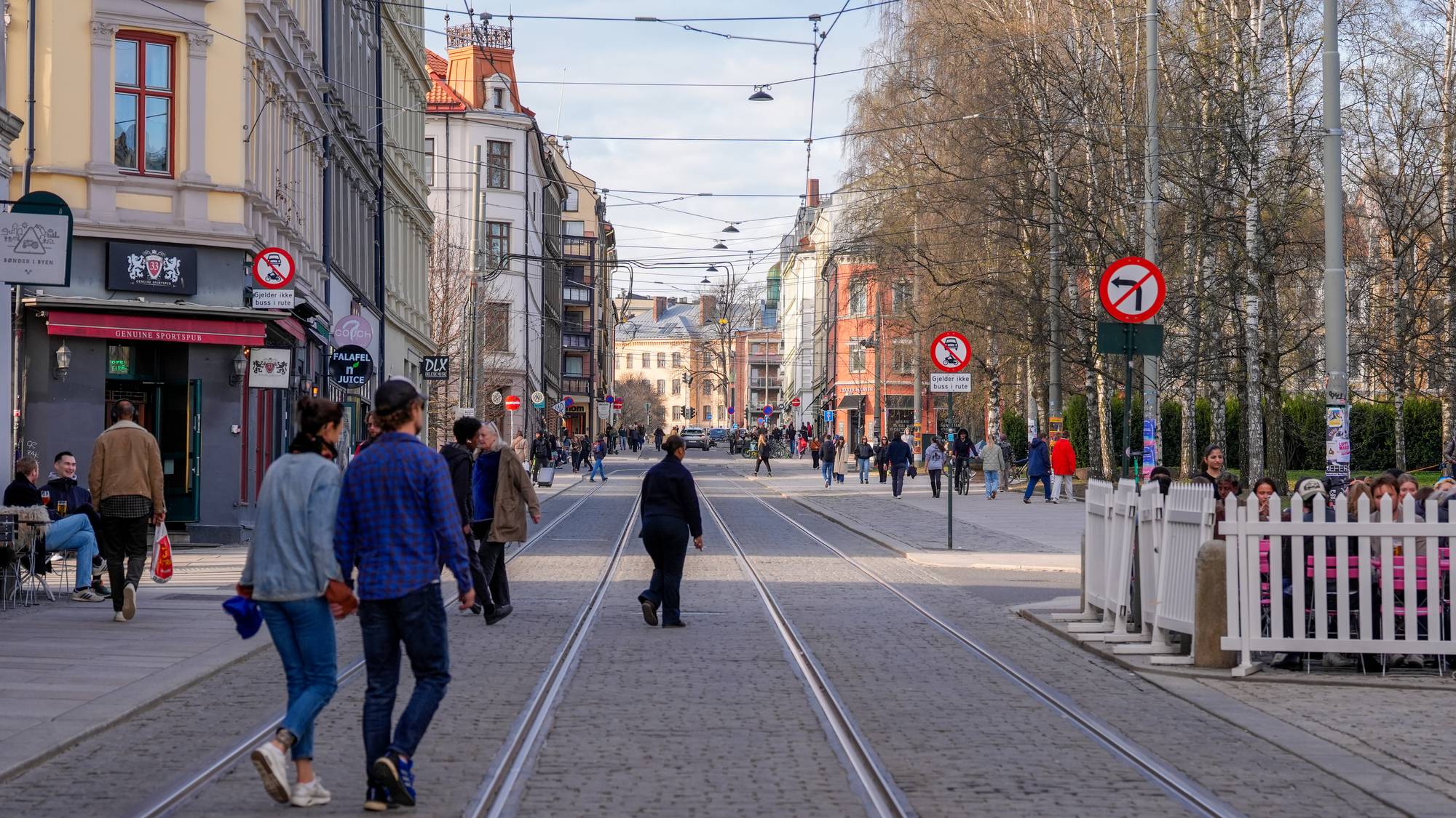 Folk går i en gate med trikkeskinner og bygårder. En uteservering er inngjerdet til høyre. (Bildebeskrivelsen er laget av en KI-tjeneste)