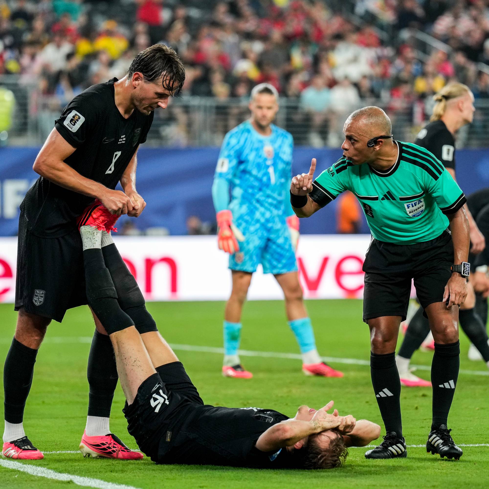 Dommeren gestikulerer med fingeren mens en fotballspiller ligger nede. En annen spiller bøyer seg for å ta av seg skoen til lagkameraten. En målvakt i blå drakt står i bakgrunnen. Publikum sitter på tribunen i en utendørs stadion. (Bildebeskrivelsen er laget av en KI-tjeneste)