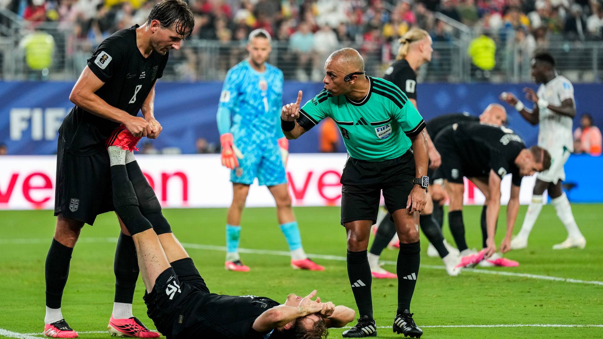 Dommeren gestikulerer med fingeren mens en fotballspiller ligger nede. En annen spiller bøyer seg for å ta av seg skoen til lagkameraten. En målvakt i blå drakt står i bakgrunnen. Publikum sitter på tribunen i en utendørs stadion. (Bildebeskrivelsen er laget av en KI-tjeneste)