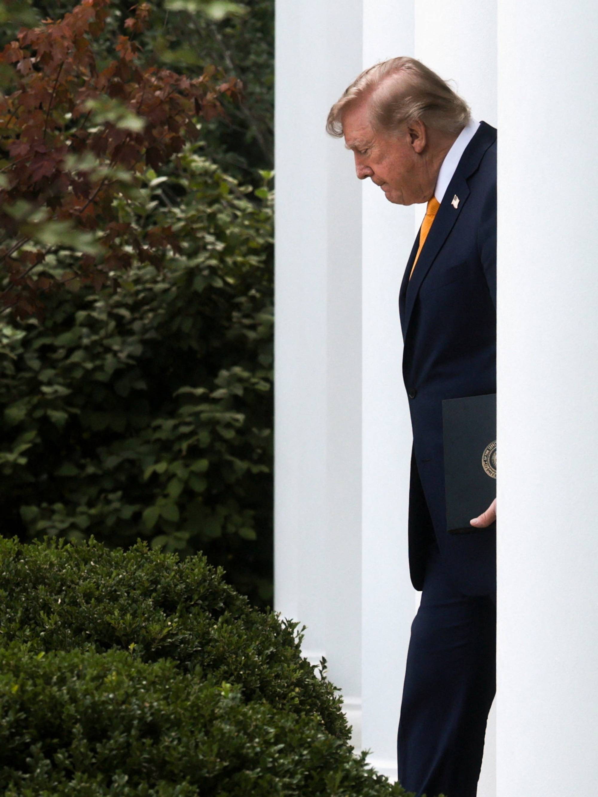 Donald Trump går ved siden av en hvit søyle med en mappe i hånden. 